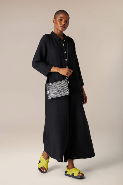 A person wearing a long black dress and bright yellow sandals stands against a neutral background, holding a Luciana Bottletop Handbag with a black and white chainmail design, handcrafted in Brazil.