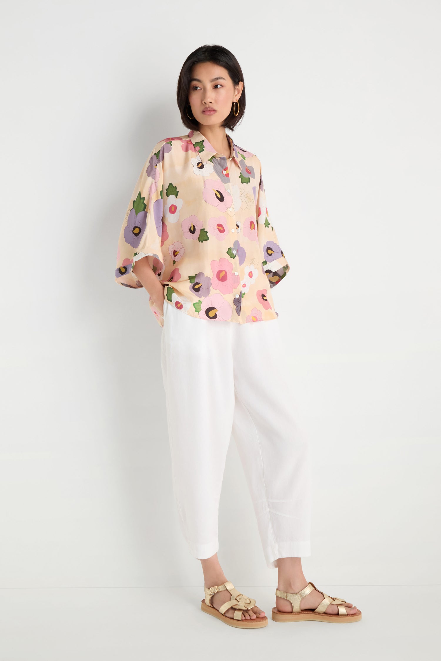 A woman stands against a white wall wearing the Rose Eclipse Silk Boxy Shirt, featuring a modern graphic floral print, paired with white loose pants and beige sandals, hands in her pockets.