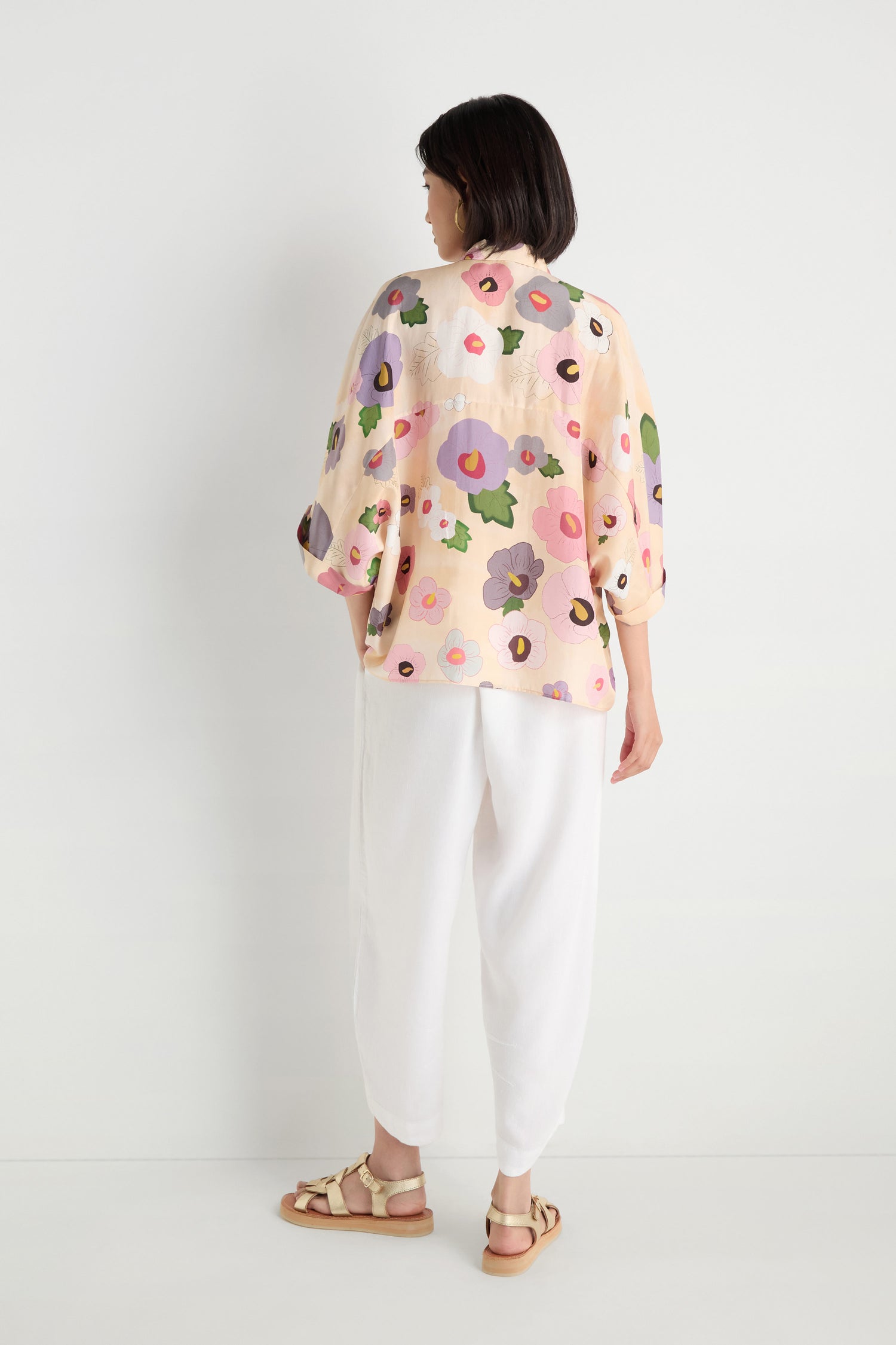 A person stands with their back to the camera against a white wall, wearing the Rose Eclipse Silk Boxy Shirt, white pants, and sandals.
