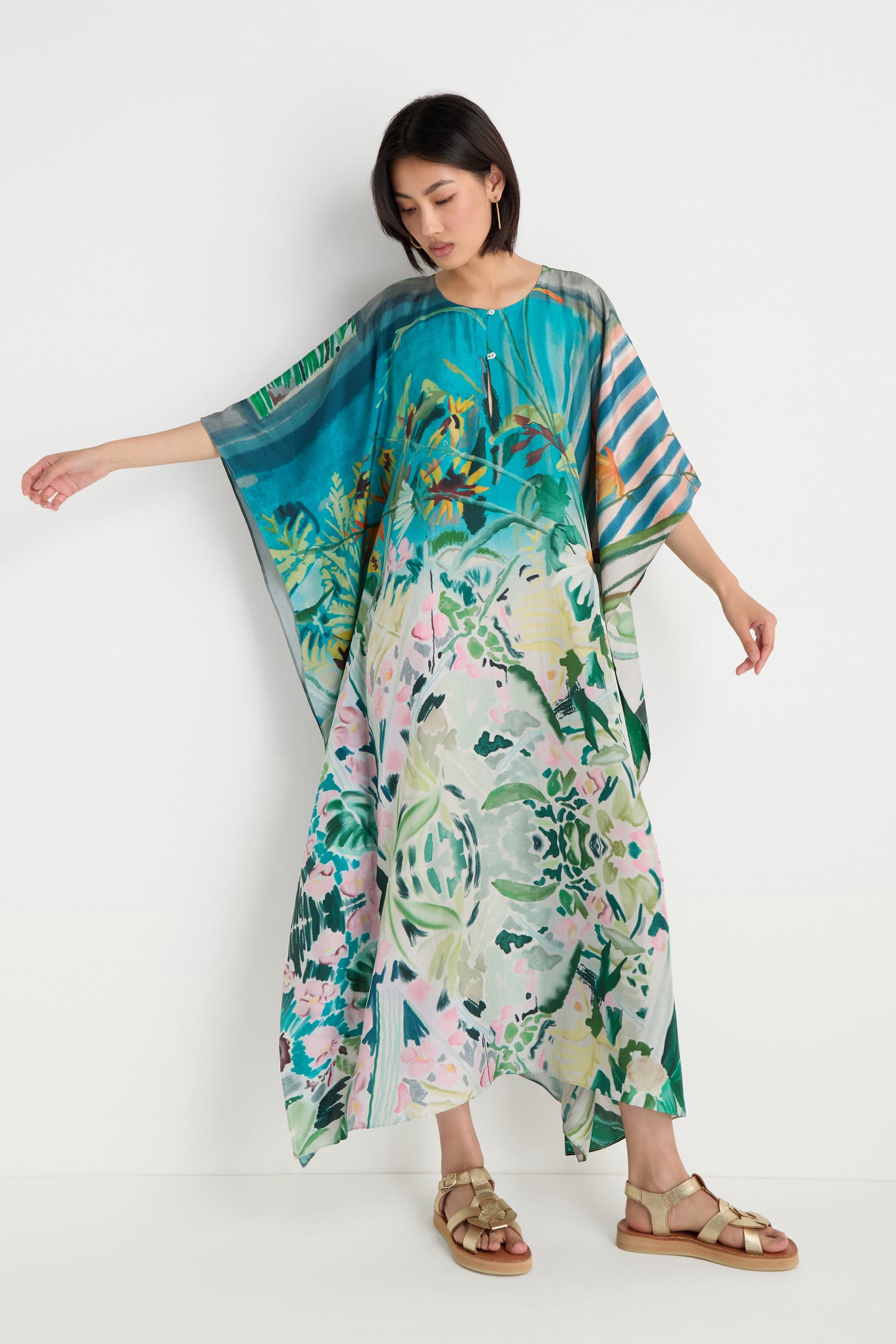 A woman stands against a white background wearing the Fern Silk Kaftan, a loose, floor-length silk dress with a colorful fern botanical print, paired with beige sandals.