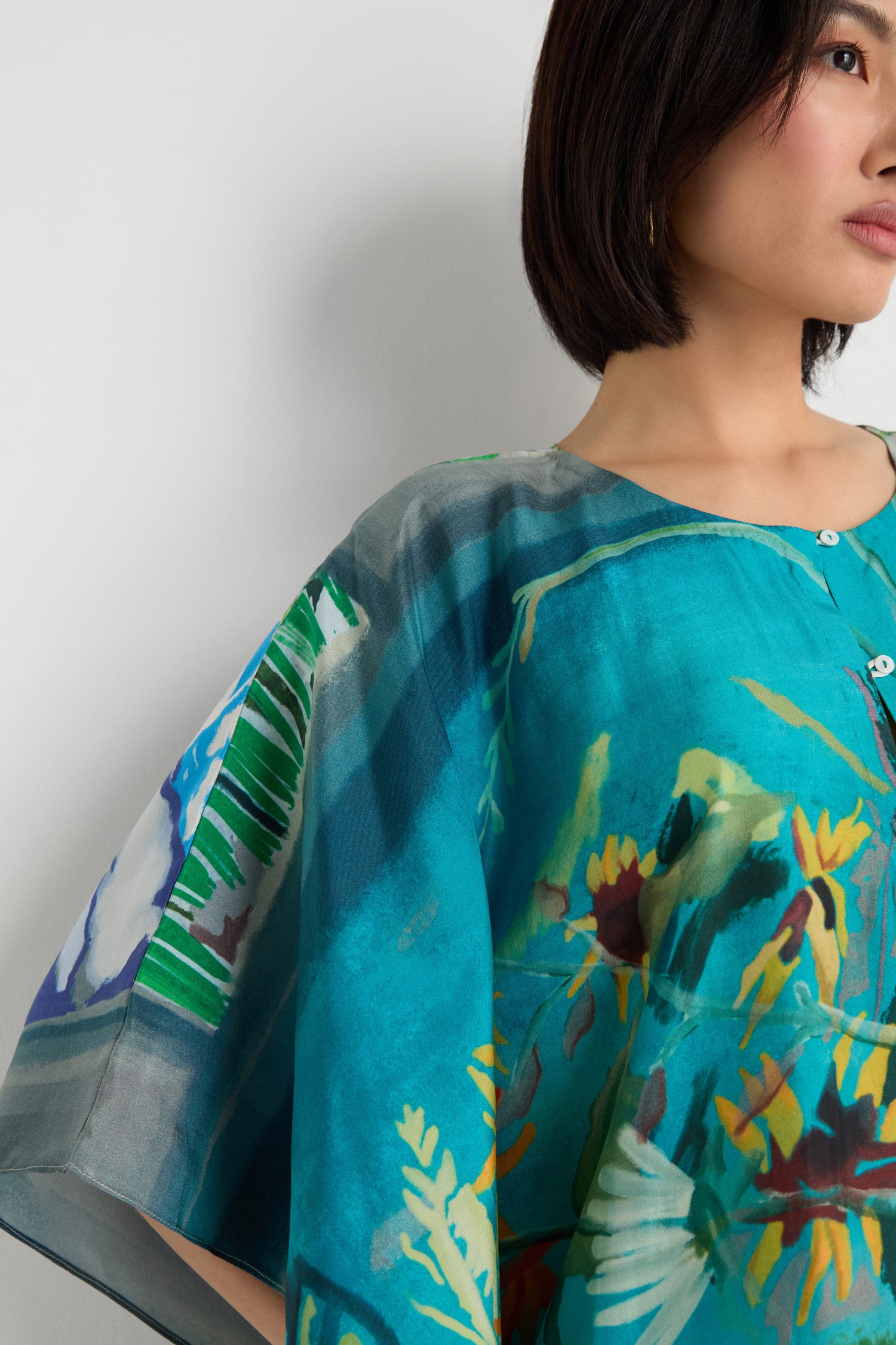 A woman with short dark hair models the Fern Silk Kaftan—a blue botanical print blouse with a flowing fit—shown from the shoulders up against a plain white background.