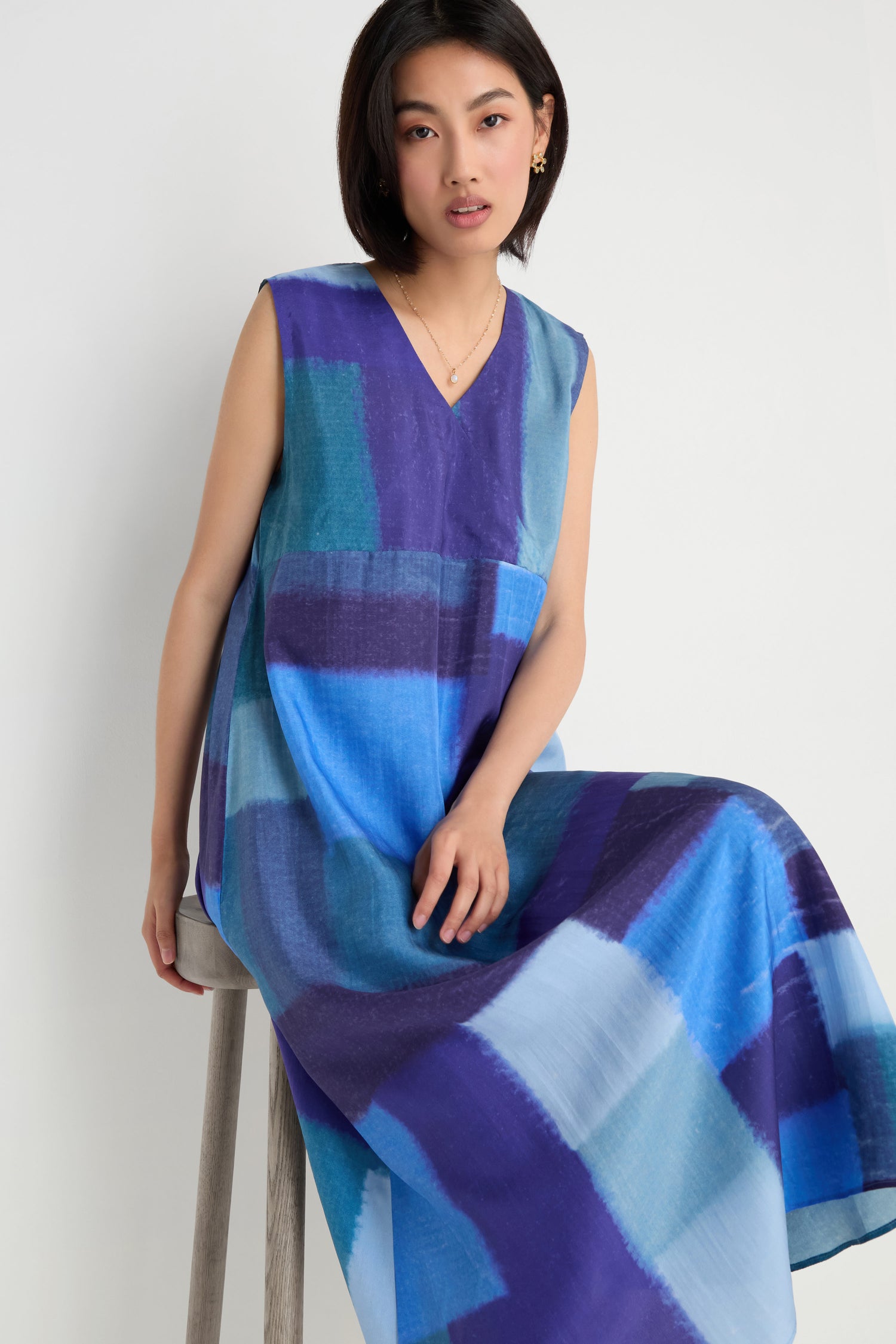 A woman in the Blurred Blocks Swing Dress, featuring sleeveless blue and purple blurred block print silk, sits on a wooden stool against a plain white background.