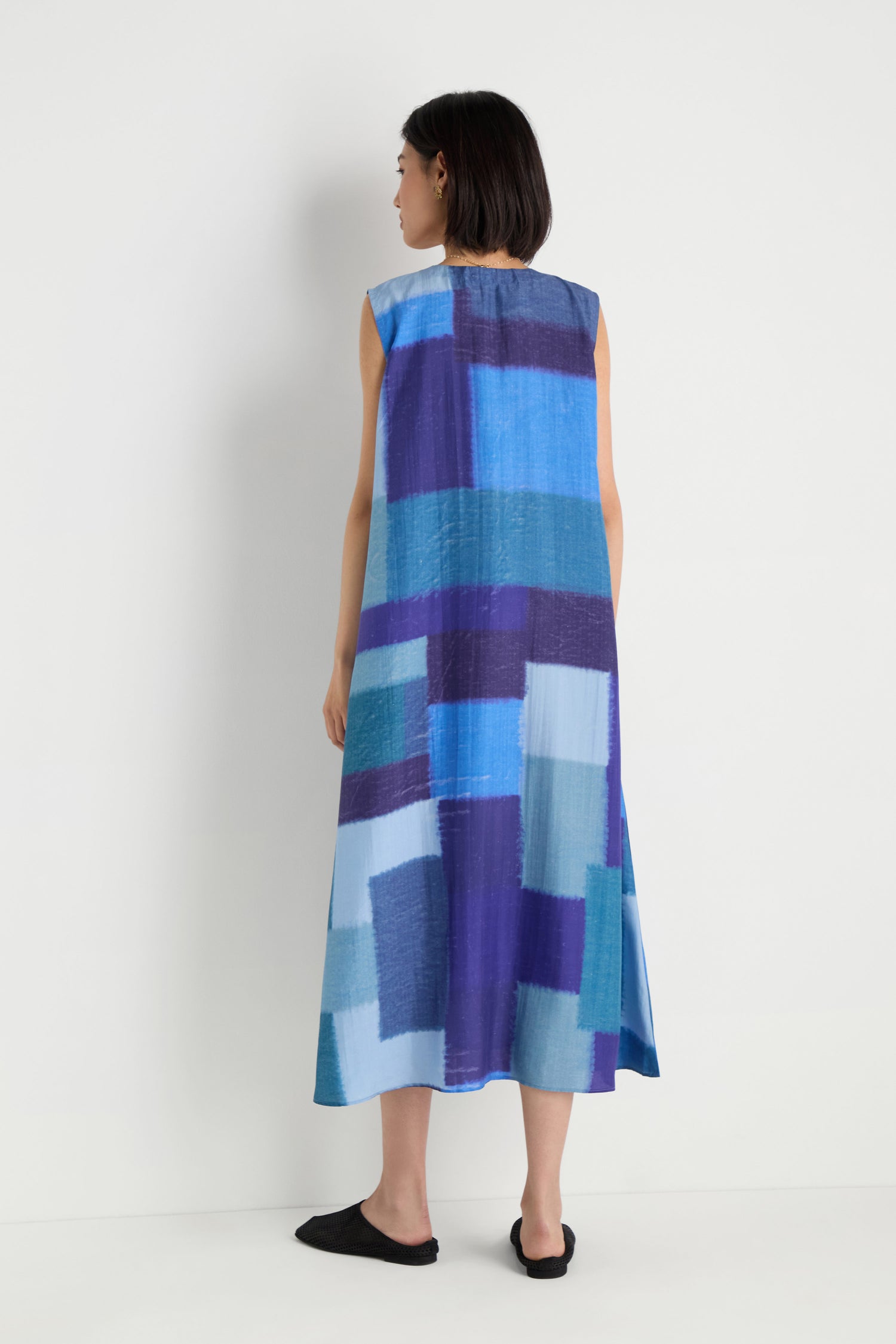 A woman faces away from the camera wearing the Blurred Blocks Swing Dress, a sleeveless silk dress featuring geometric blue and purple patterns, set against a plain white background.