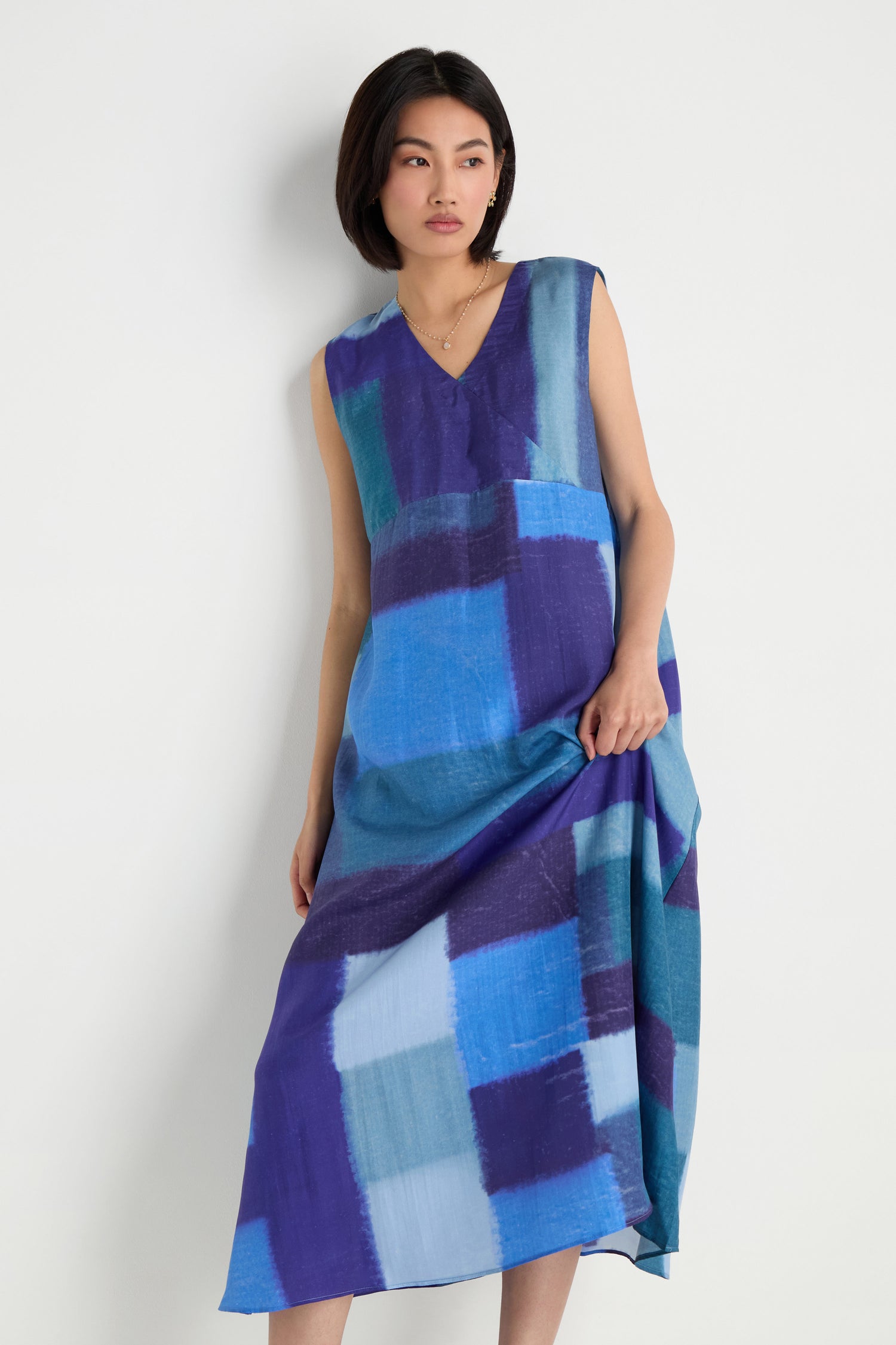 A woman models the Blurred Blocks Swing Dress—a sleeveless, loose-fitting midi dress with a blue blurred block print—while standing against a plain white background and looking slightly to the side.