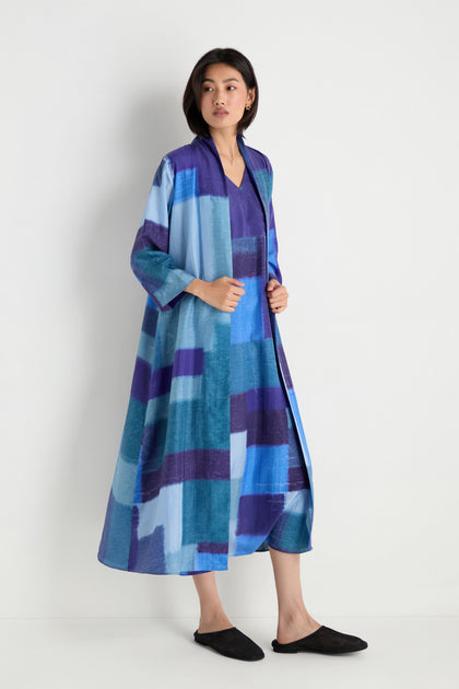 A woman stands against a white background wearing the Blurred Blocks Easy Jacket—a long, loose-fit piece with blue and purple geometric patterns—styled as a lightweight layer and paired with black slip-on shoes.