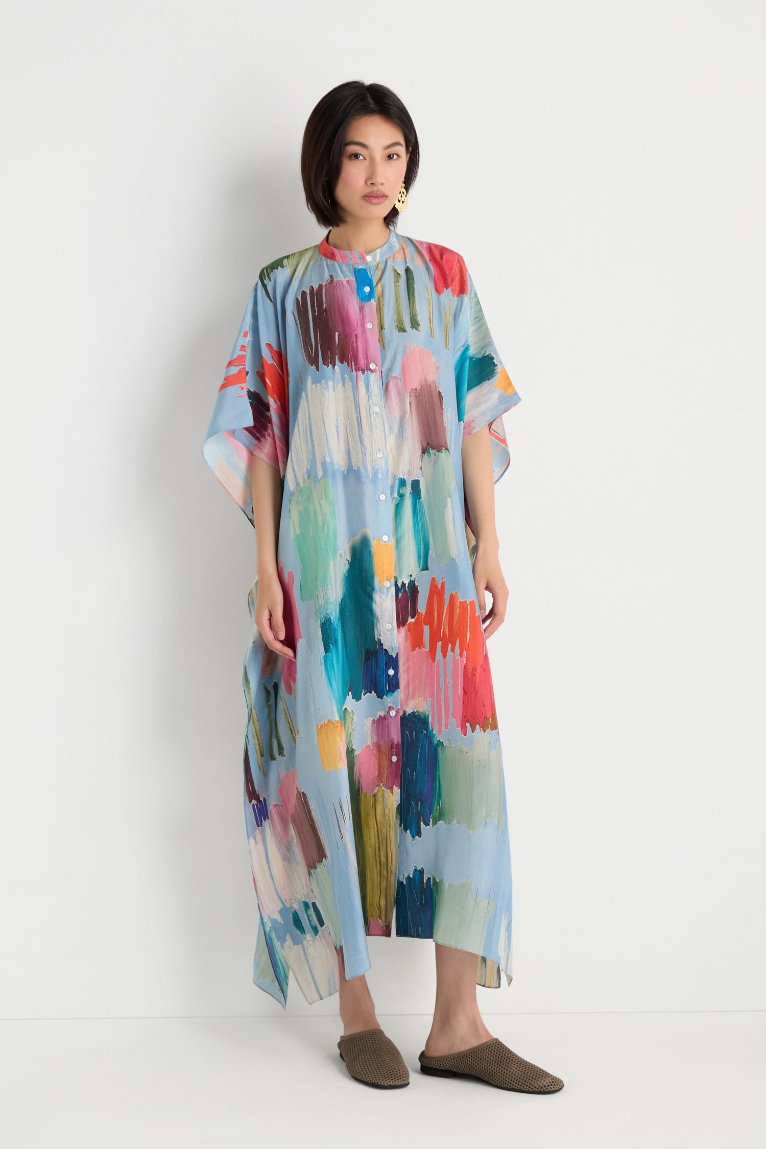 A woman stands against a white wall wearing the Breezy Strokes Silk Shirt Dress, a loose maxi with a colorful brushstroke print, paired with brown woven slip-on shoes.