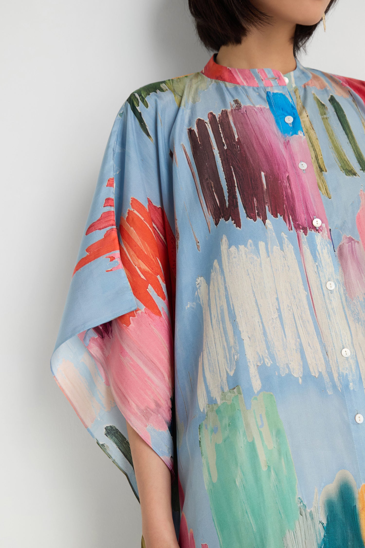 A person wearing the Breezy Strokes Silk Shirt Dress, a loose button-up design with wide sleeves and a colorful brushstroke print on a light blue background.