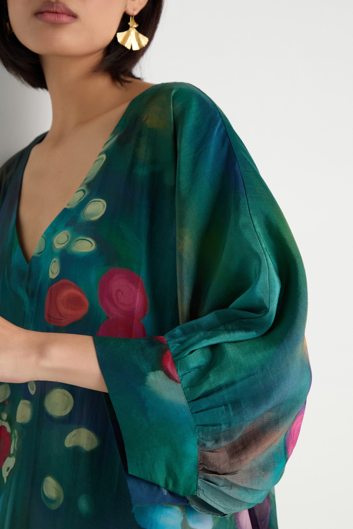 Woman wearing the Soft Floral Silk V-Neck Dress in a green and blue abstract floral print with loose sleeves, paired with gold geometric earrings, shown from shoulders to mid-arm.