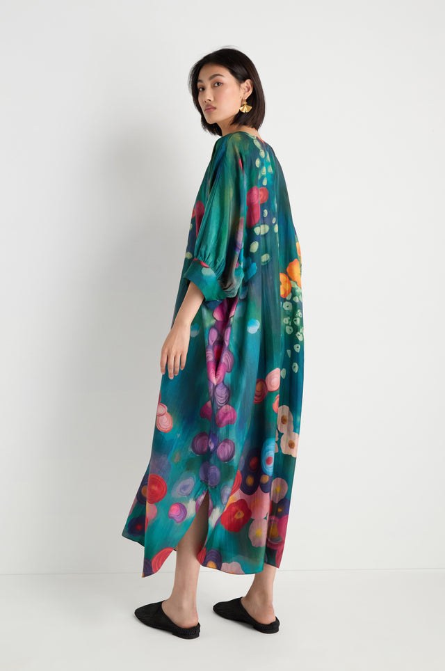 A woman stands sideways against a plain white background, wearing the Soft Floral Silk V-Neck Dress with an abstract floral print, paired with black flats and gold earrings.