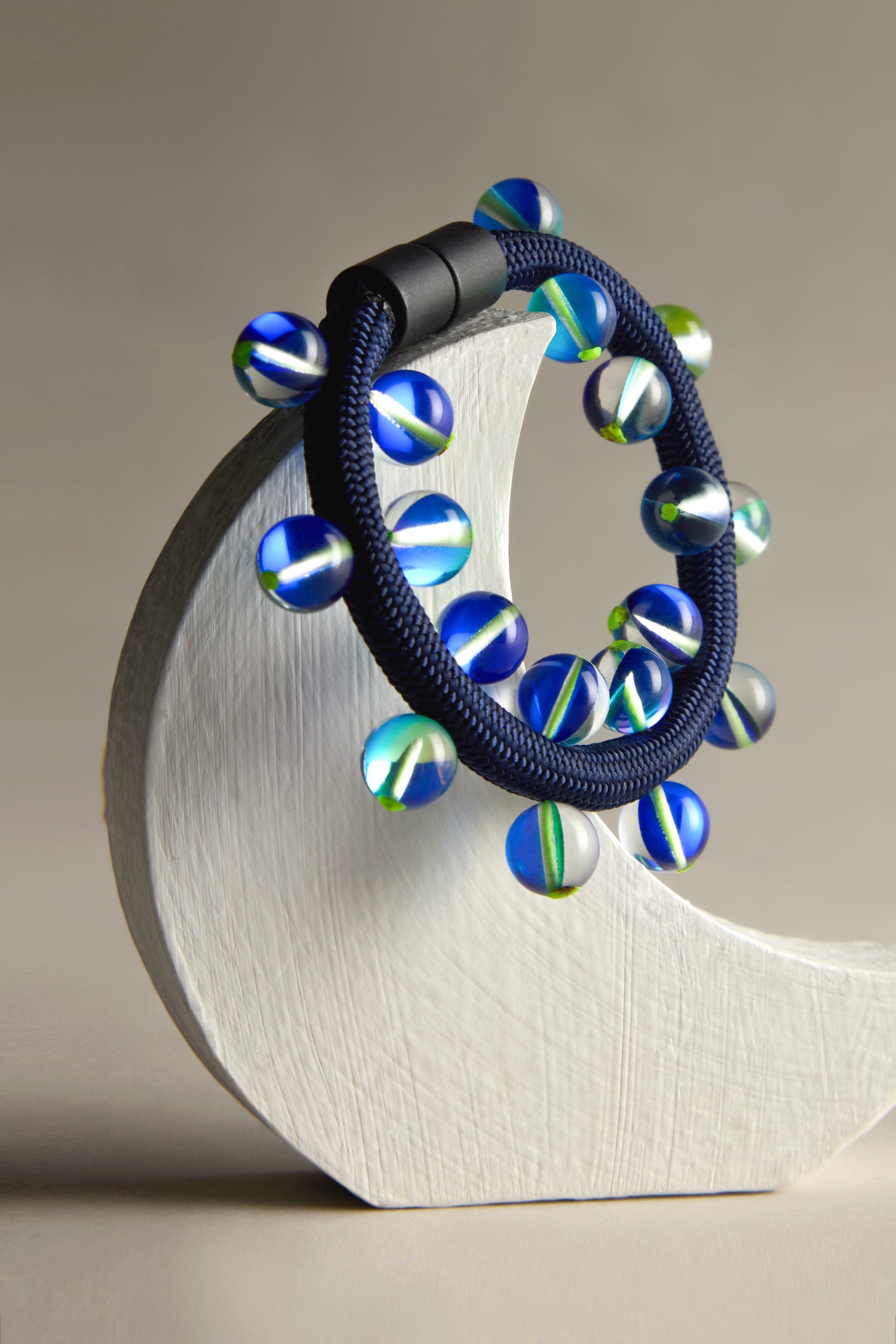 The Christina Brampti Atmosphere Bracelet is a modern jewelry piece with round blue and green beads on a blue-and-black band, beautifully showcased on a white crescent stand against a neutral backdrop.