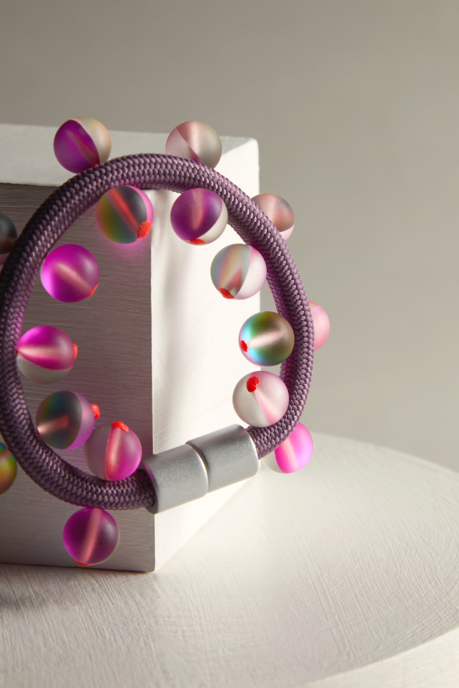 The Atmosphere Bracelet by Christina Brampti showcases a sleek design with purple and multicolored translucent beads, artfully presented on a white block against a neutral setting.