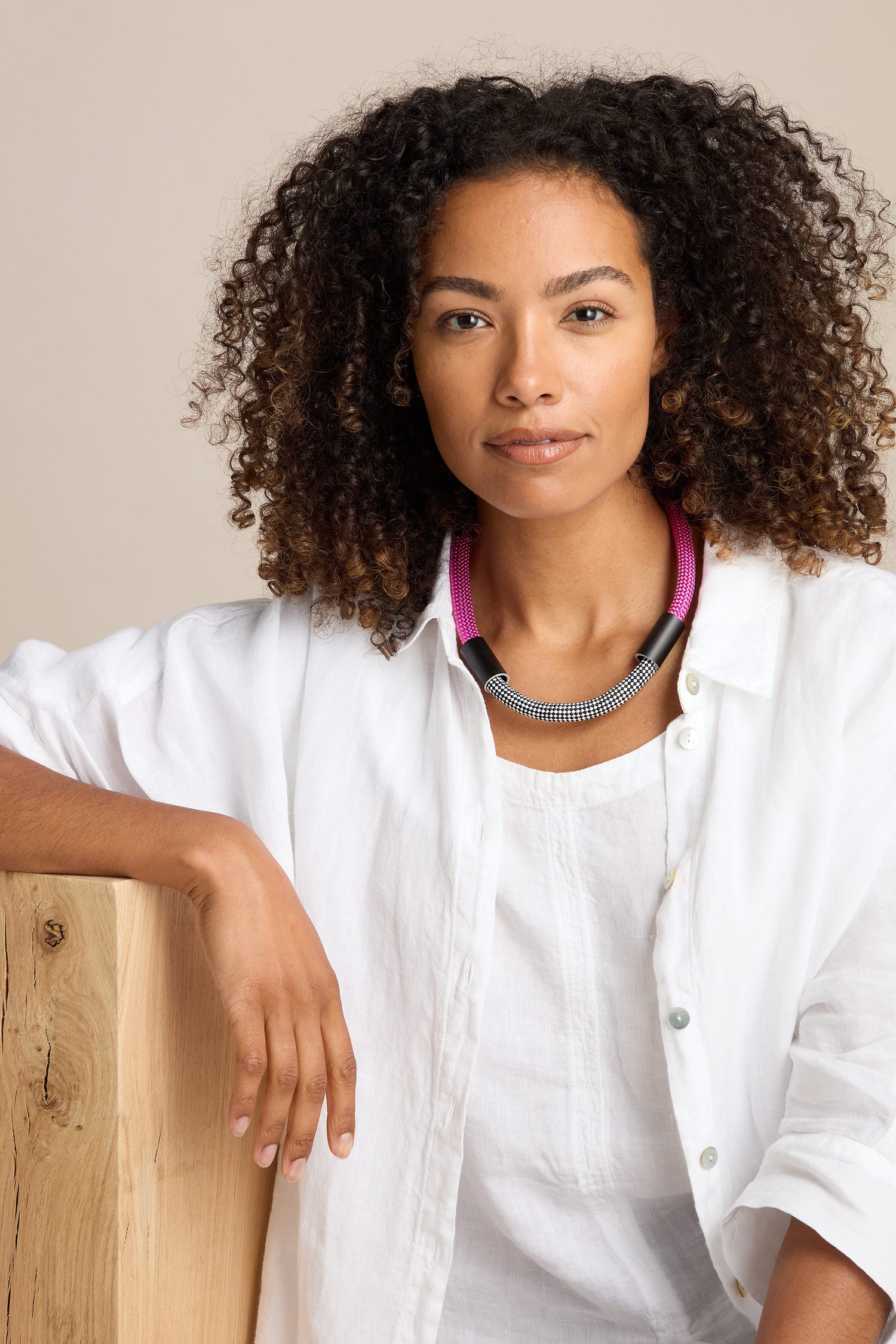 A person with curly hair wearing a white shirt and a modern statement piece—the Short Cord Necklace in pink and black—rests one arm on a wooden surface.