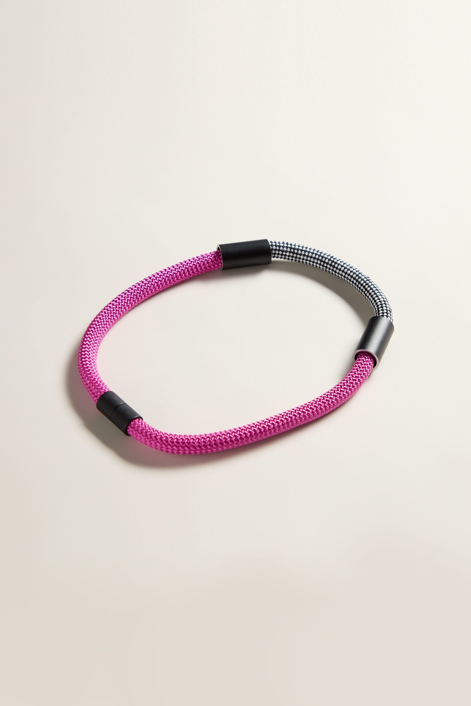 Introducing the Short Cord Necklace: A stylish necklace crafted with a braided cord featuring a pink section and a black-and-white checkered section, connected by sleek black plastic clasps. This modern accessory is perfect for making a statement.