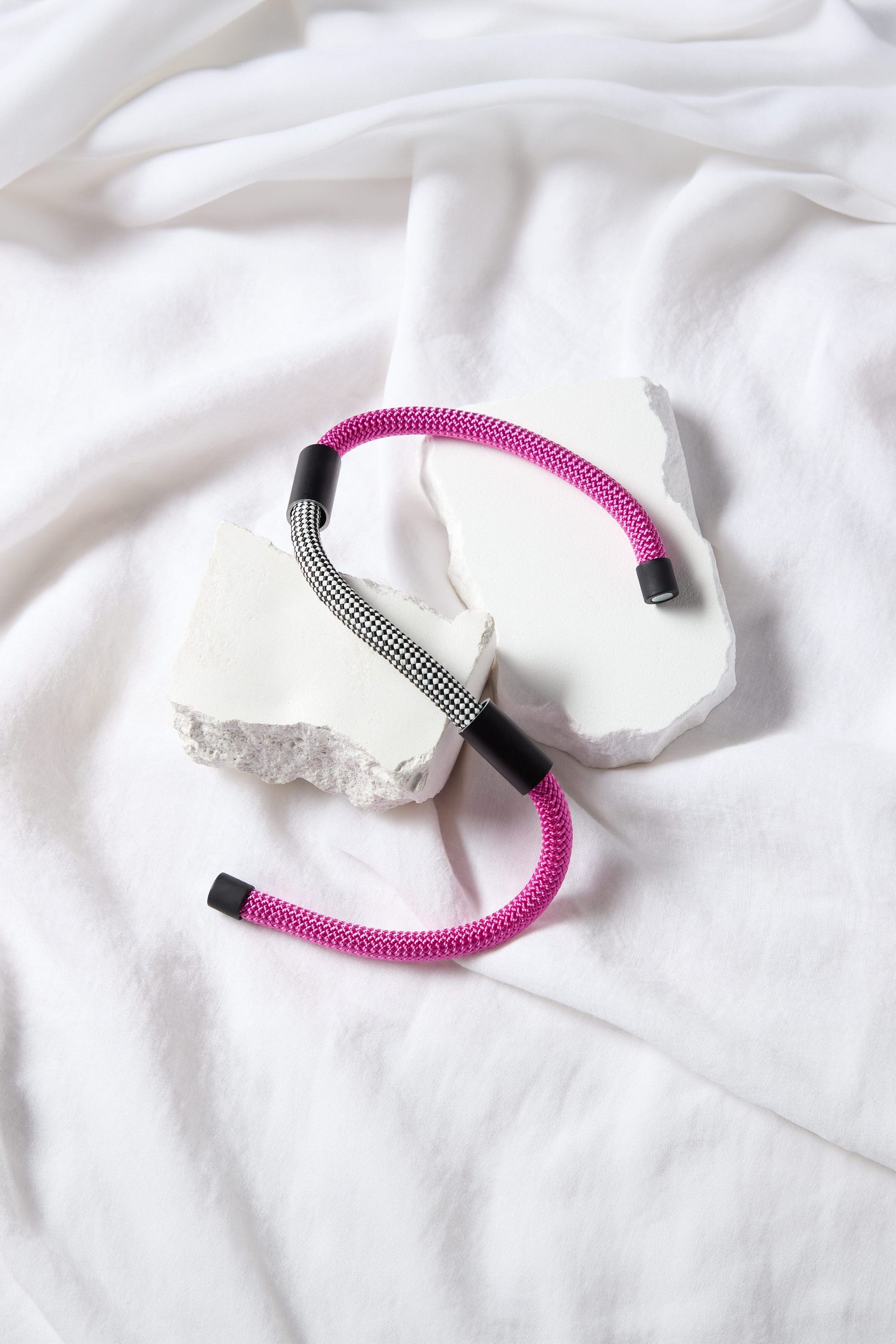 The Short Cord Necklace, featuring two braided cables—one in pink and the other in black and white—set against broken white blocks and a white fabric background, creates a striking statement piece perfect as a modern accessory or stylish necklace.