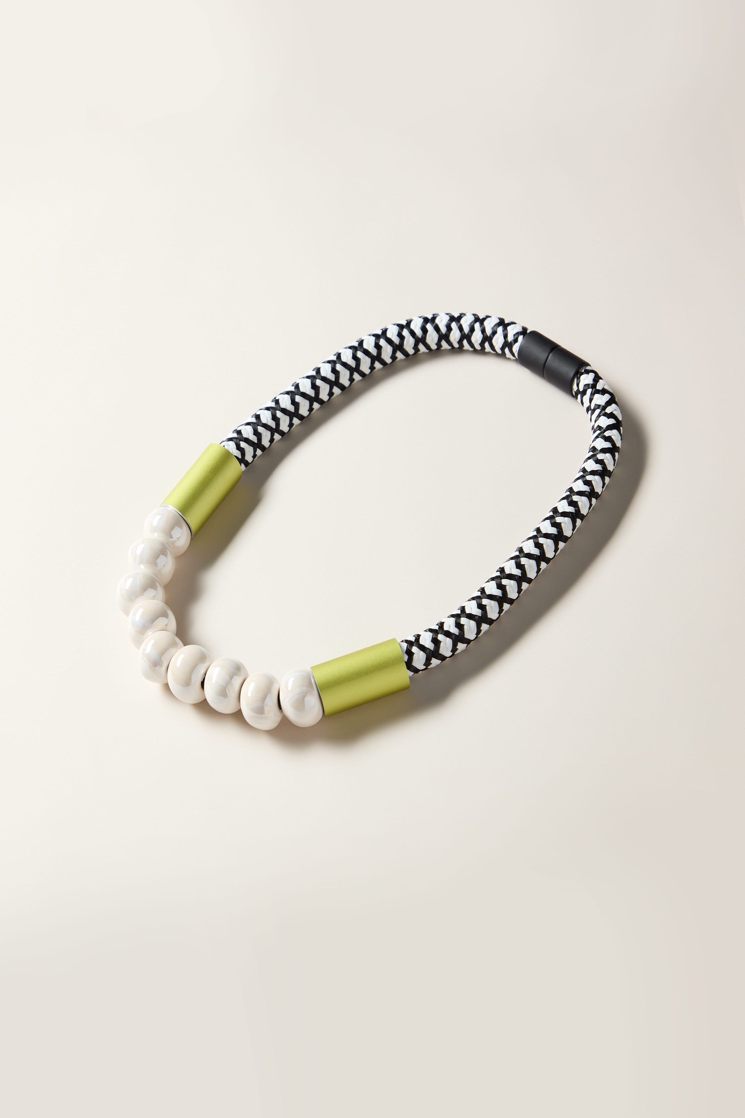 The Milky Way Pearlescent Necklace, featuring a braided black and white design accented with green end caps and pearlescent ceramic beads, is displayed on a light background, presenting a contemporary style for those in search of a bold statement piece.