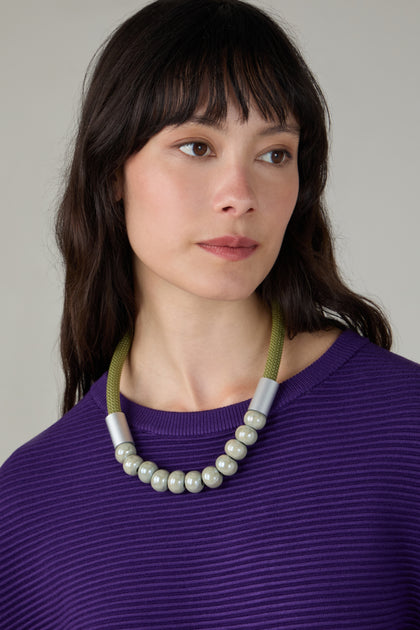 A woman with long dark hair, wearing a purple ribbed top and the Olive Ceramic Milkyway Necklace, featuring large metallic beads, looks slightly to the side against a neutral background.