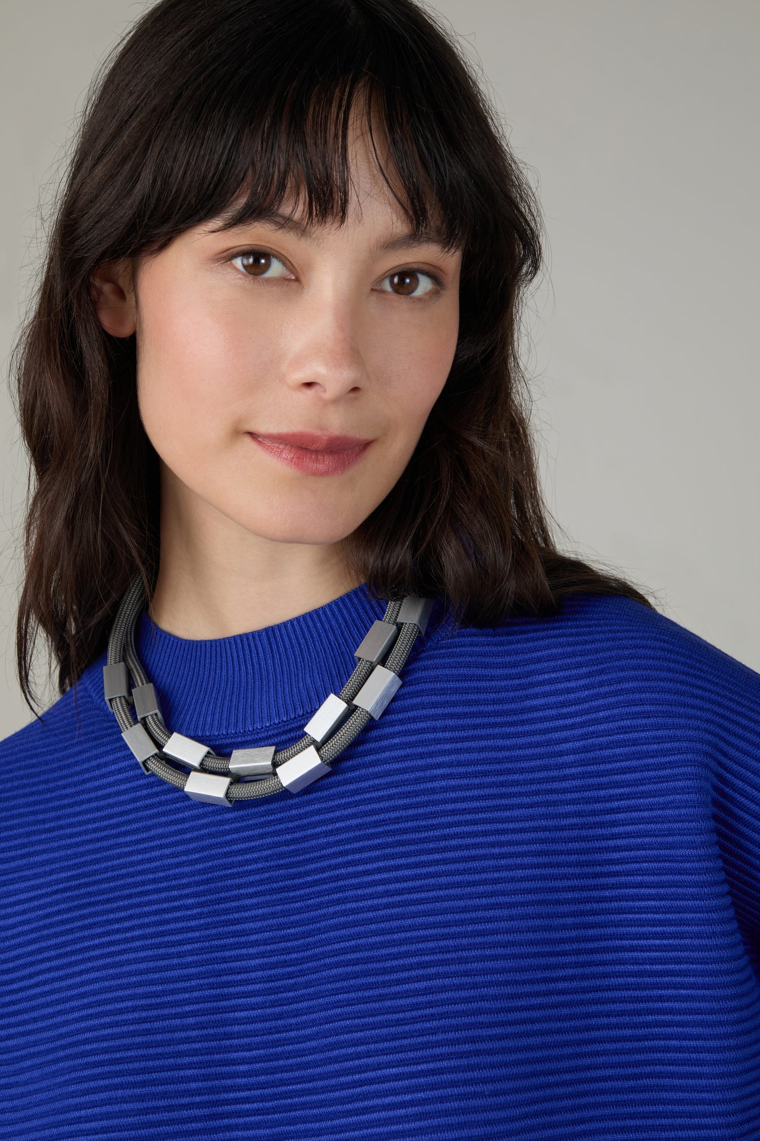 A woman with medium-length dark hair wears a cobalt blue ribbed sweater and the Short Aluminium Cube Cord Necklace, posing against a neutral background.