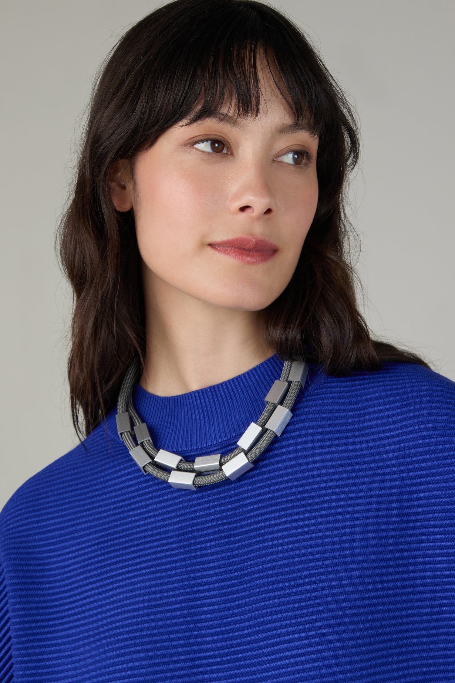 A woman with long dark hair, wearing a bright blue ribbed top and the Short Aluminium Cube Cord Necklace, looks slightly to the side against a neutral background.