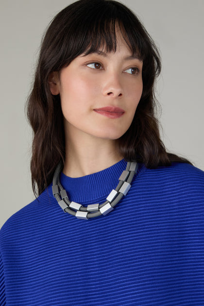 A woman with long dark hair, wearing a bright blue ribbed top and the Short Aluminium Cube Cord Necklace, looks slightly to the side against a neutral background.