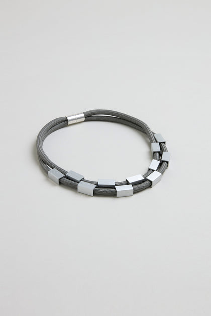 The Short Aluminium Cube Cord Necklace features gray multi-strand cords with evenly spaced aluminum cube beads and a cylindrical clasp, shown on a light gray background.