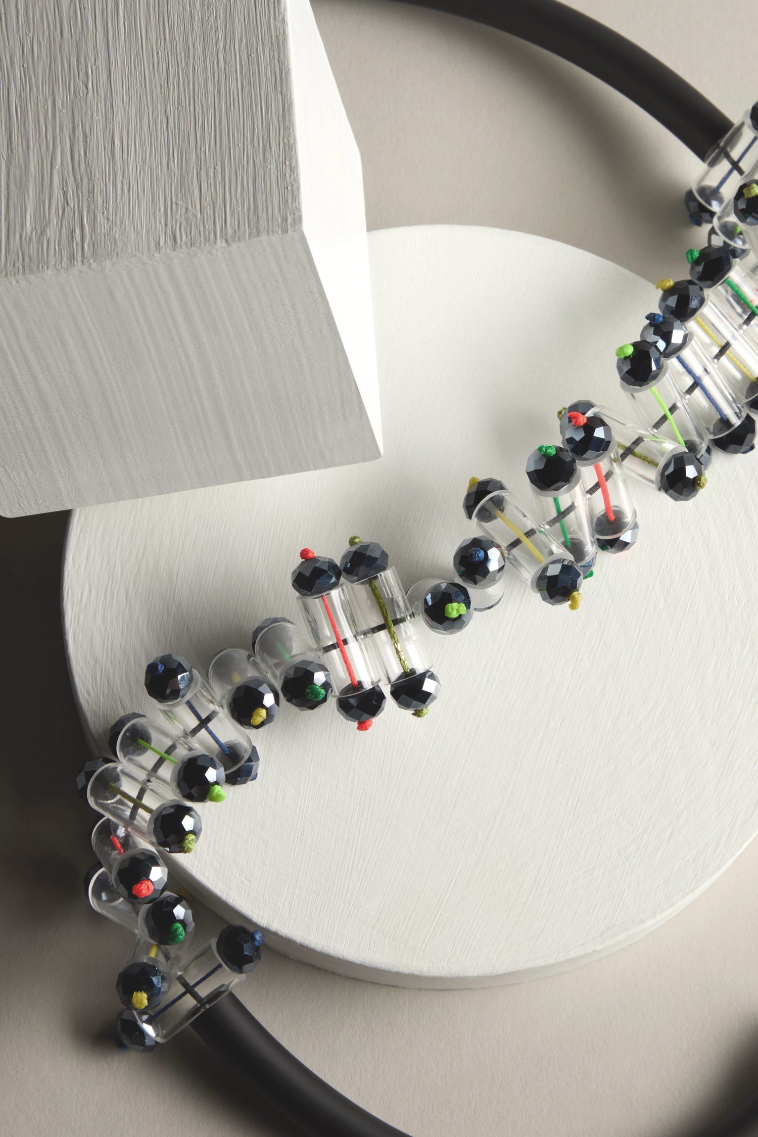 A Matrix Necklace model with colored connectors and black atoms rests on a circular platform resembling a futuristic necklace, accompanied by a rectangular block against a soft gray background.