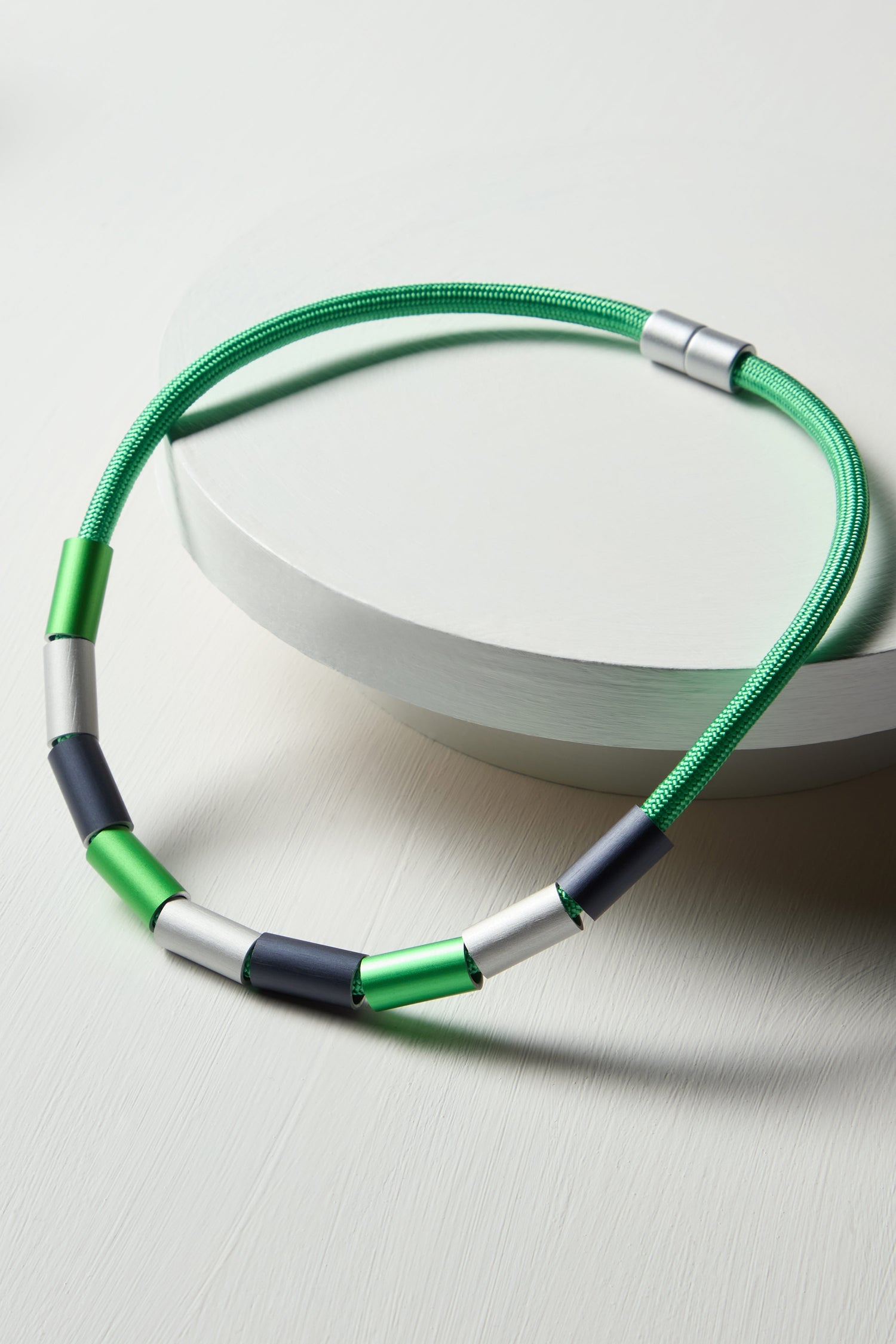 A green and black cord bracelet with silver cylindrical beads, evoking the sleek design of Christina Brampti's Aluminium Cylinders Necklace, elegantly displayed on a curved, light gray surface.