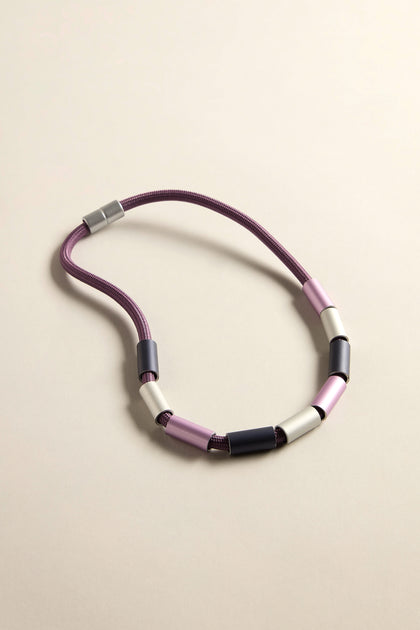 Explore the Aluminium Cylinders Necklace, a statement piece with a modern touch. It features a purple cord adorned with alternating pink, black, and silver cylindrical beads, elegantly arranged in a loop against a neutral backdrop.