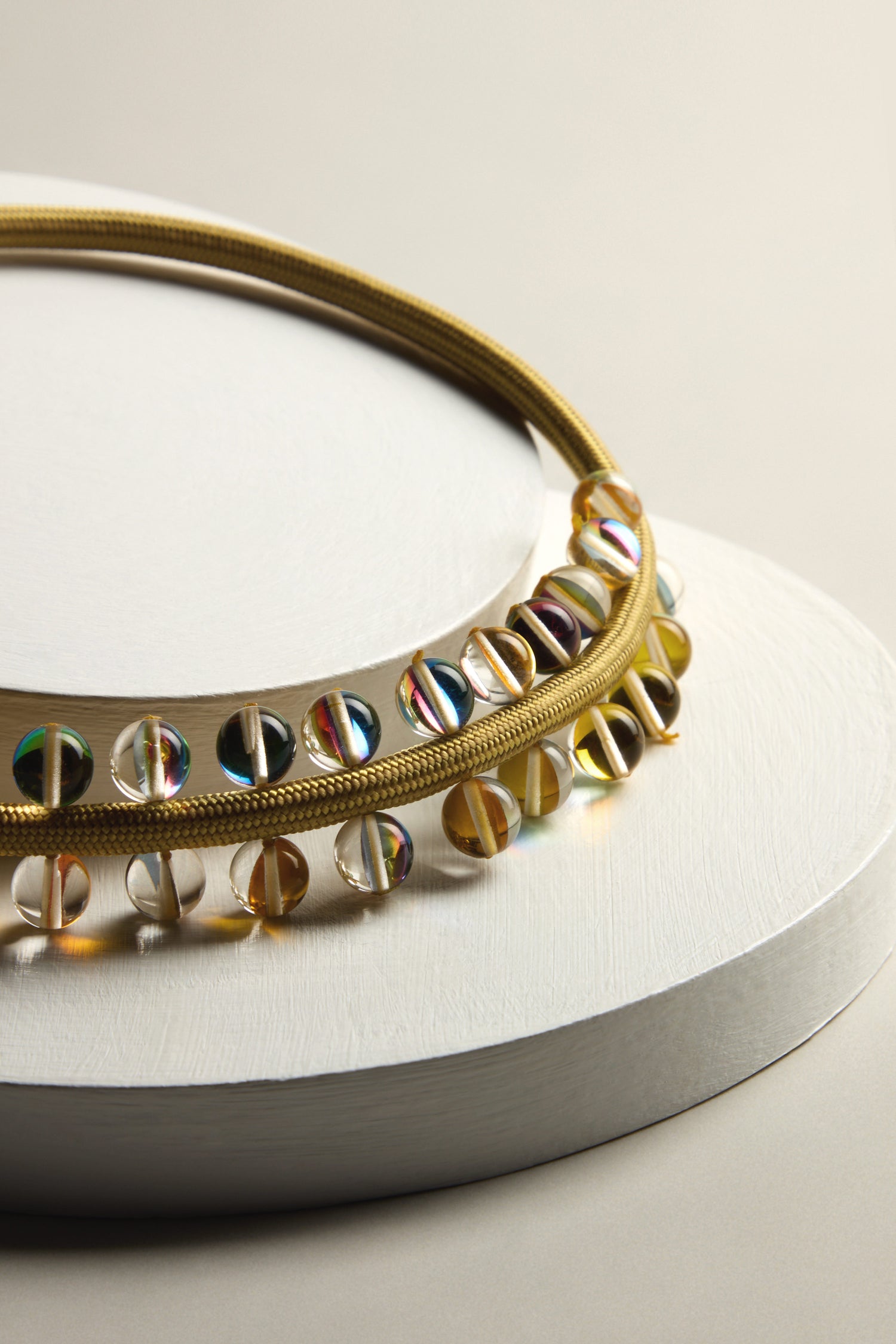 The Atmosphere Necklace, with its iridescent beads, is gracefully displayed on a white textured circular platform set against a neutral backdrop.