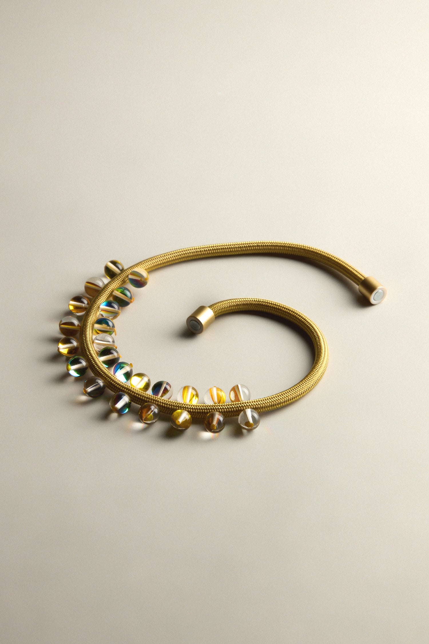 Introducing the Atmosphere Necklace: a coiled gold work of art with shimmering beads on one side, elegantly contrasted against a simple light backdrop. It captures modern design, infusing elegance and color into any outfit.