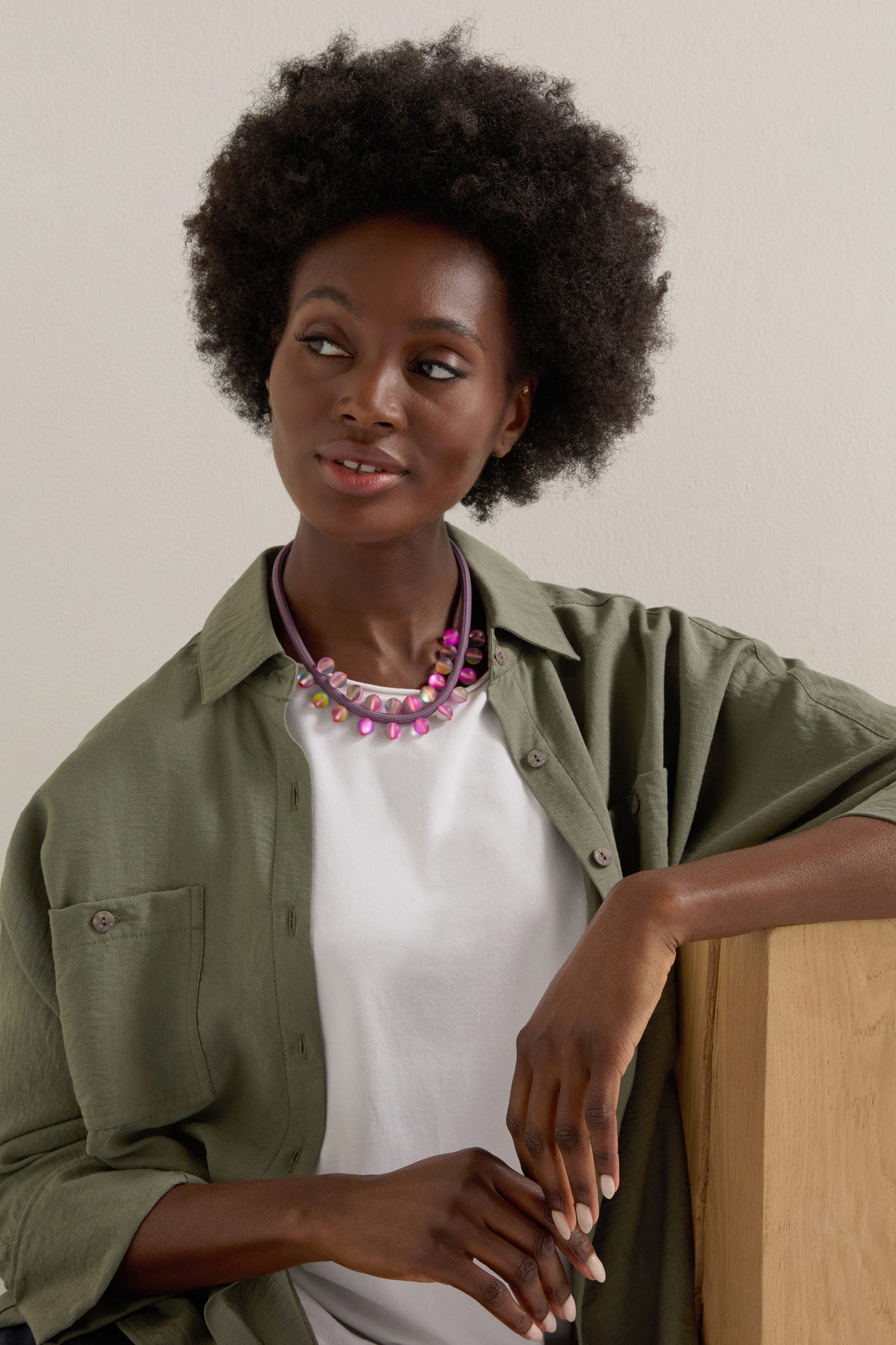 Sporting an afro hairstyle, a person in a green jacket and white shirt leans on wood while showcasing the pink Atmosphere Necklace, perfectly blending modern artistry with chic elegance.