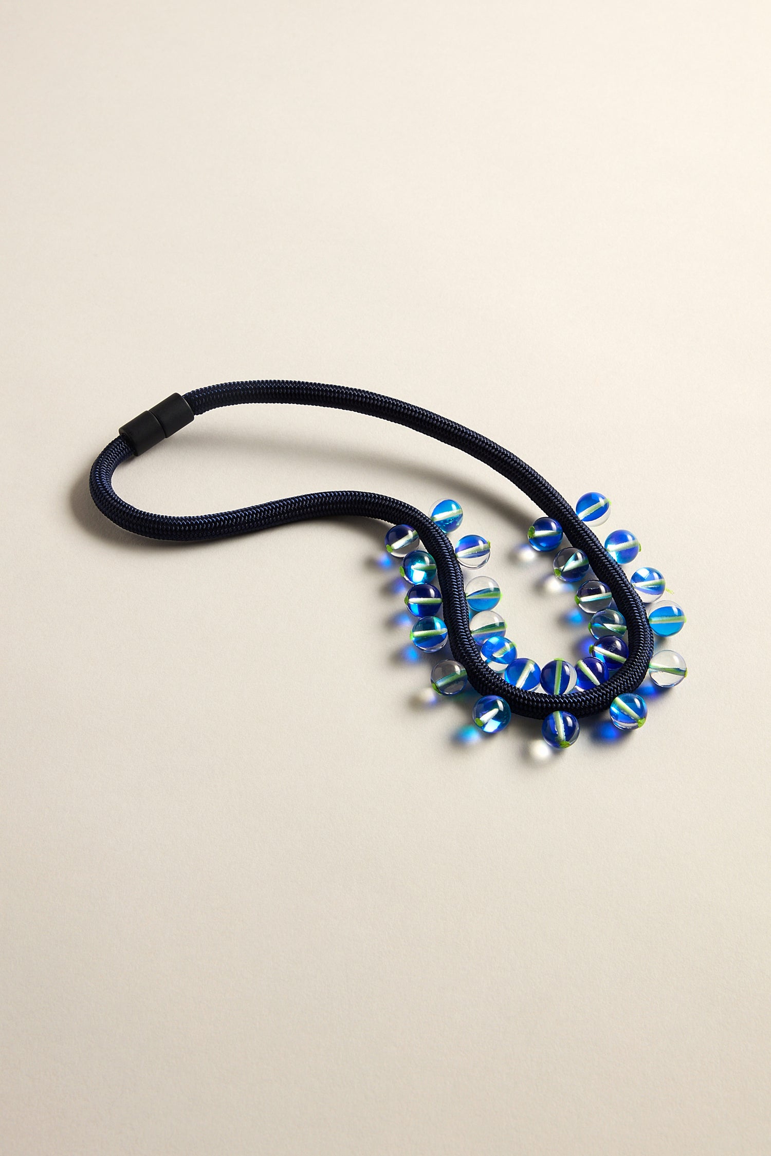 Discover the Atmosphere Necklace, a dark blue rope with a modern artistic flair. It showcases iridescent blue and green beads in diverse shapes on a light backdrop.