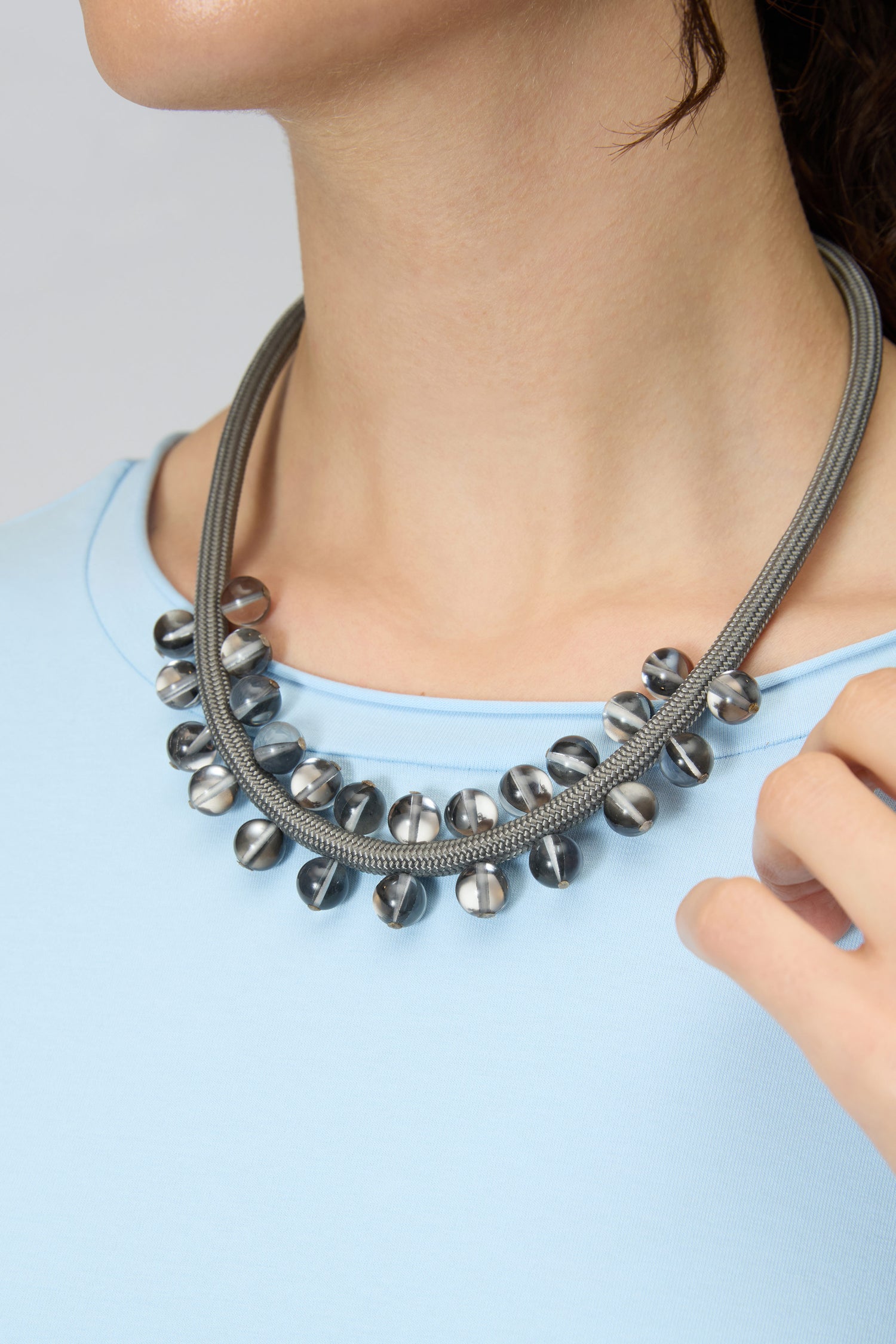 A person wearing the Smoke Atmosphere Necklace, a double-stranded gray contemporary piece with metallic and glass beads, paired with a light blue top.
