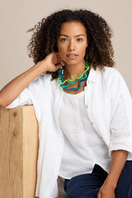 A person with curly hair wears a white shirt and a vibrant Cord Waves Necklace, sits next to a wooden surface, rests their head on their hand, and looks at the camera.