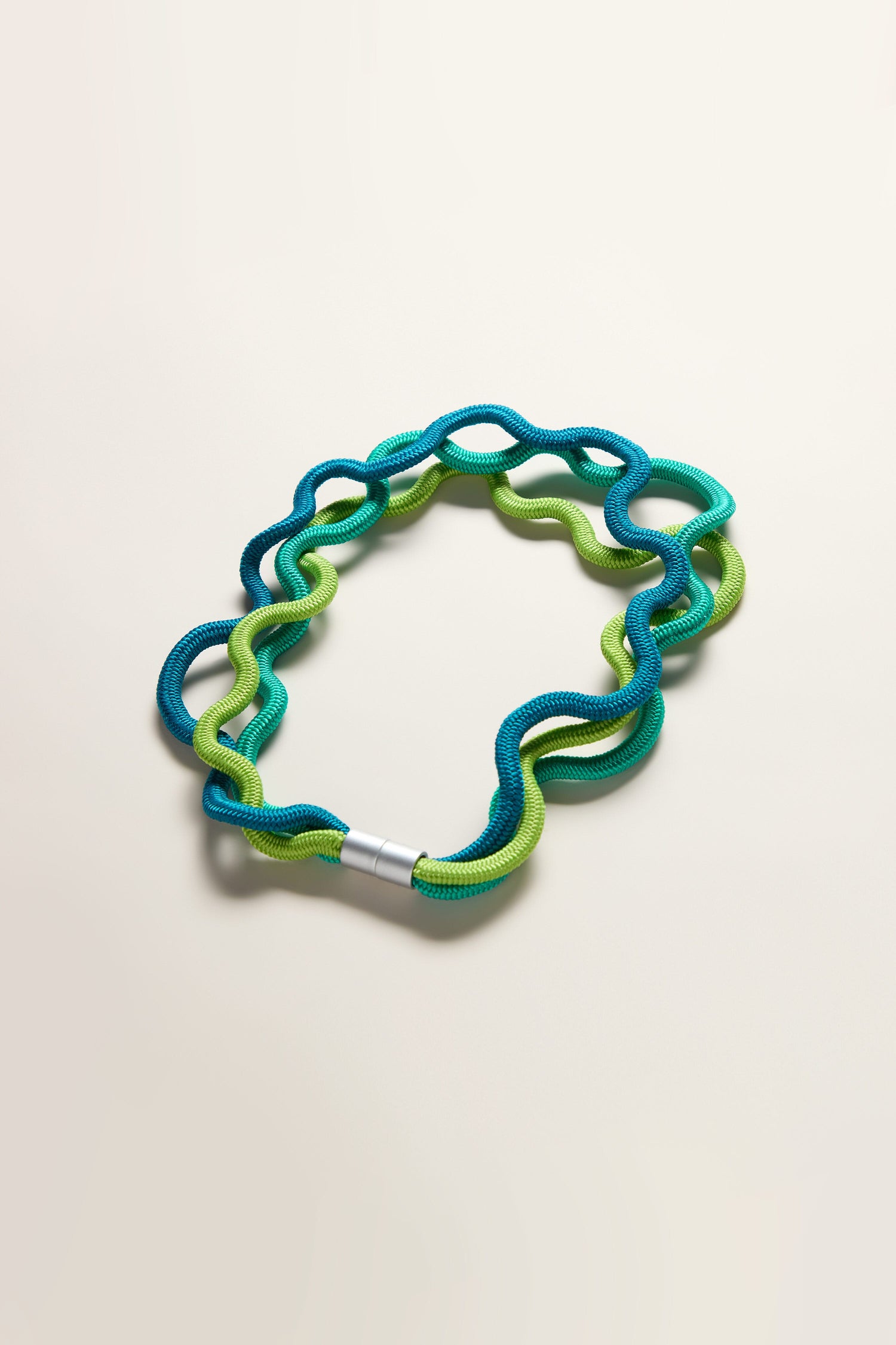 A vibrant blue and green flexible twistable toy with a silver connector, fashioned into a loop on a neutral background, reminiscent of the modern flair found in Christina Brampti's Cord Waves Necklace.