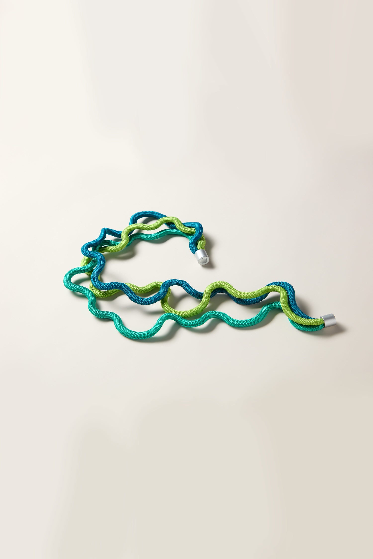 A Cord Waves Necklace, featuring coiled green and blue bungee cord with metal hooks on both ends and resembling a piece of contemporary jewellery, is laid out on a plain light background.