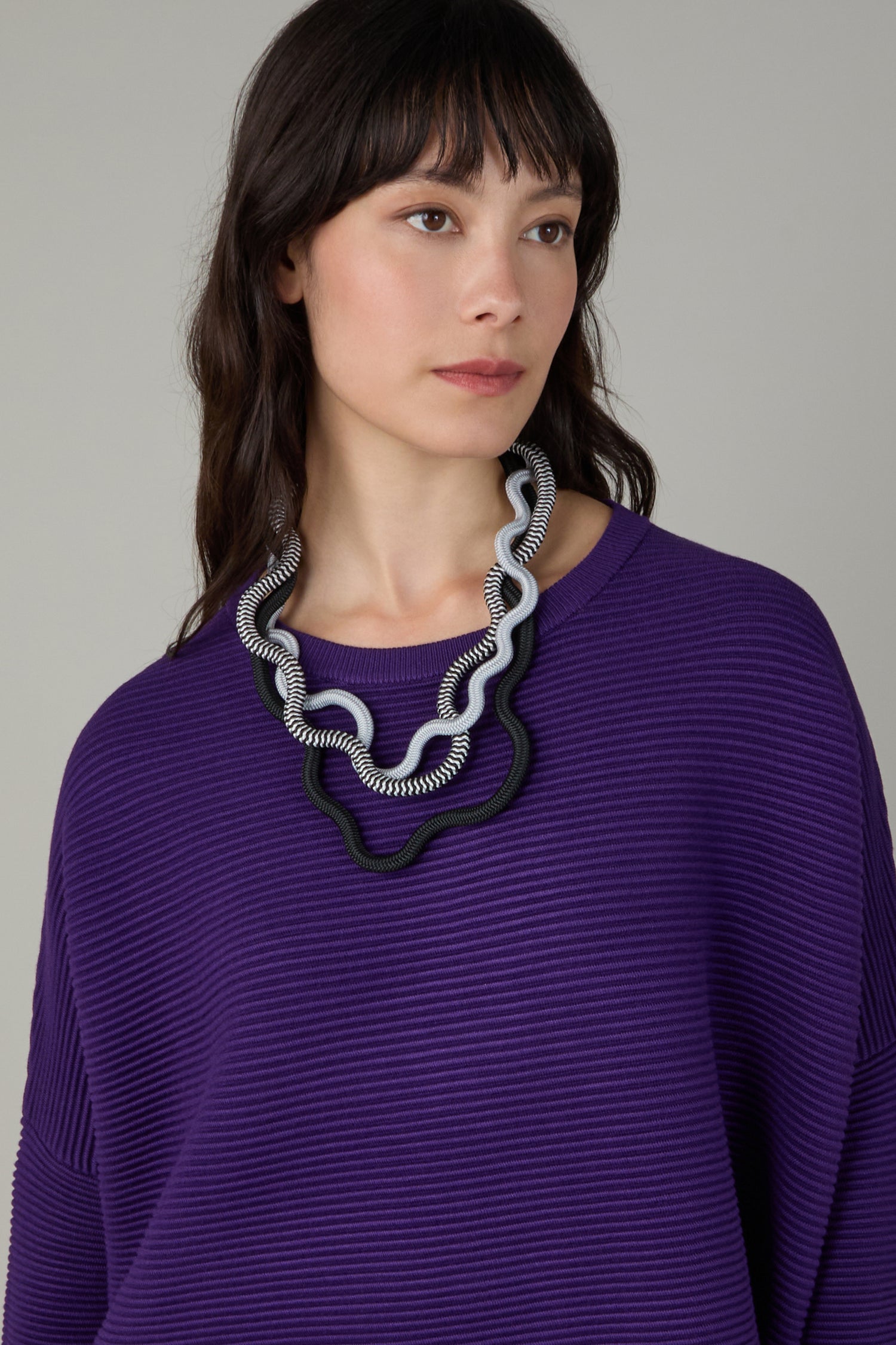 Woman wearing a ribbed purple top and the Black and White Monochrome Cord Waves Necklace by Christina Brampti, looking slightly to the side against a plain background.