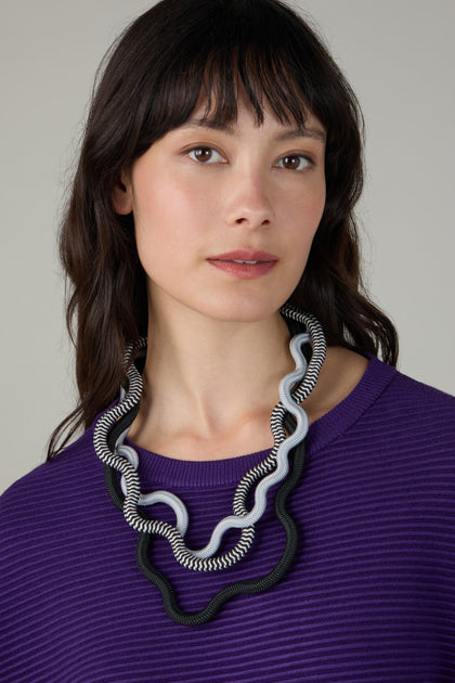 A woman with long dark hair, wearing a purple ribbed top and the Black and White Monochrome Cord Waves Necklace by Christina Brampti, looks at the camera.