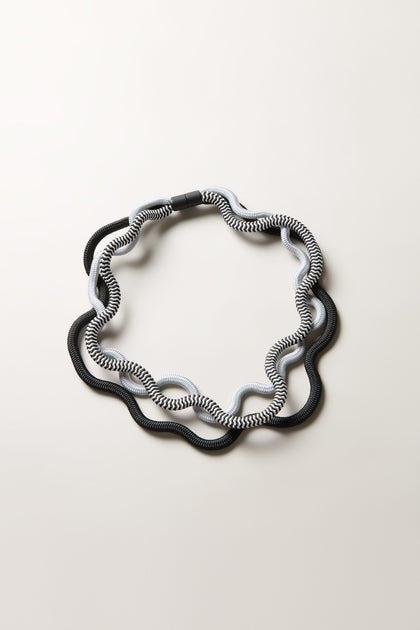 The Black and White Monochrome Cord Waves Necklace is laid flat on a light surface, its wavy circular form and alternating black, white, and gray segments resembling a sculptural jewelry piece or modern monochrome necklace.