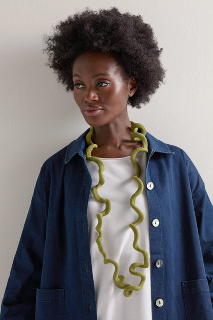 A person wearing a Long Twist Cord Necklace by Christina Brampti is styled over a white shirt and denim jacket, standing against a light-colored wall.