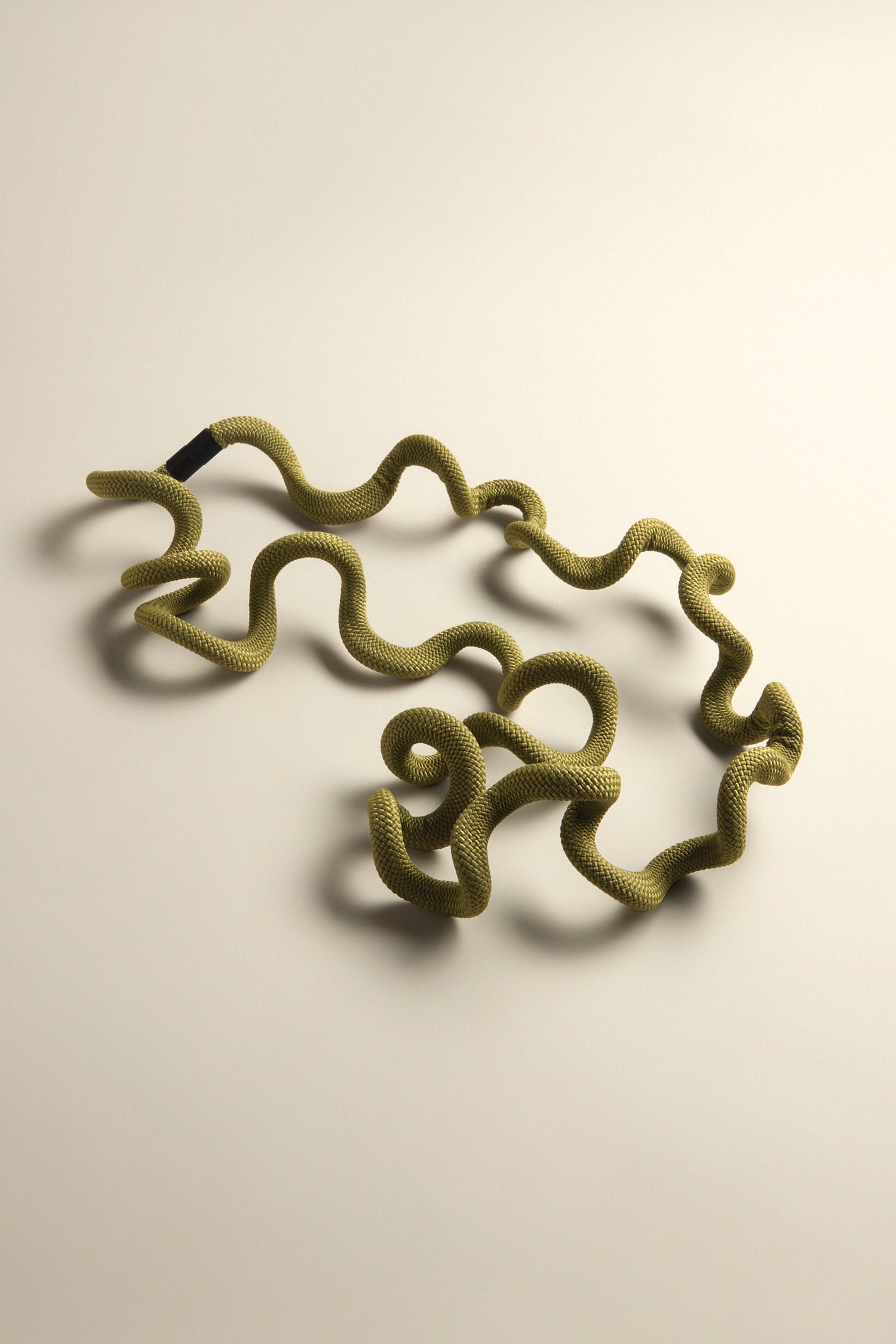 A Long Twist Cord Necklace in olive green with small black accents, styled like a Christina Brampti piece, is laid casually on a beige surface.