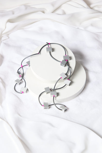 A contemporary statement piece, the Long Aluminium Cube Necklace by Chrisina Brampti showcases geometric silver beads with pink accents, artfully arranged on white circular platforms set against a backdrop of crumpled white fabric.