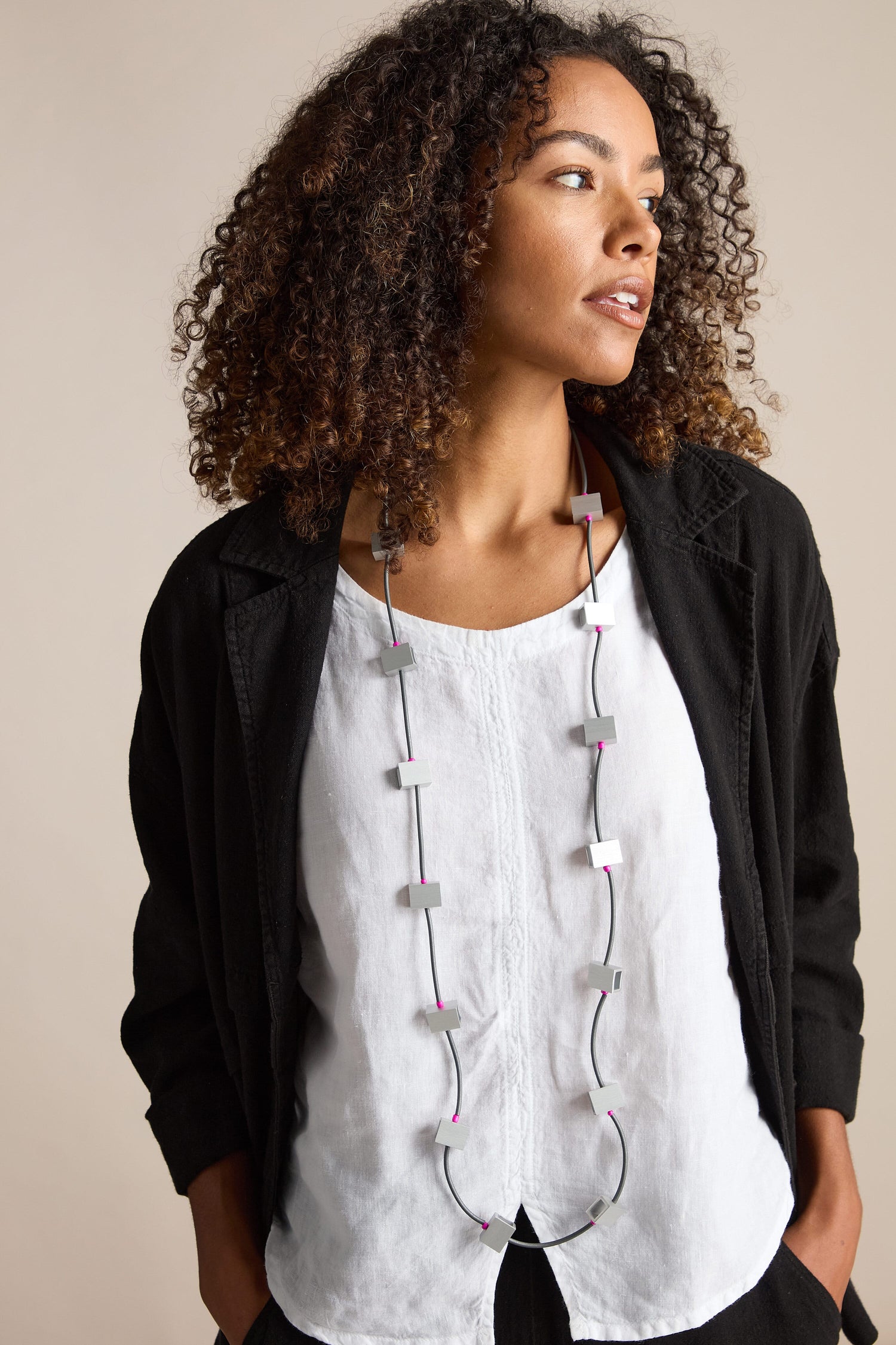 A person with curly hair looks to the side, wearing a white shirt, black jacket, and a Long Aluminum Cube Necklace by Chrisina Brampti, making it a bold statement piece.