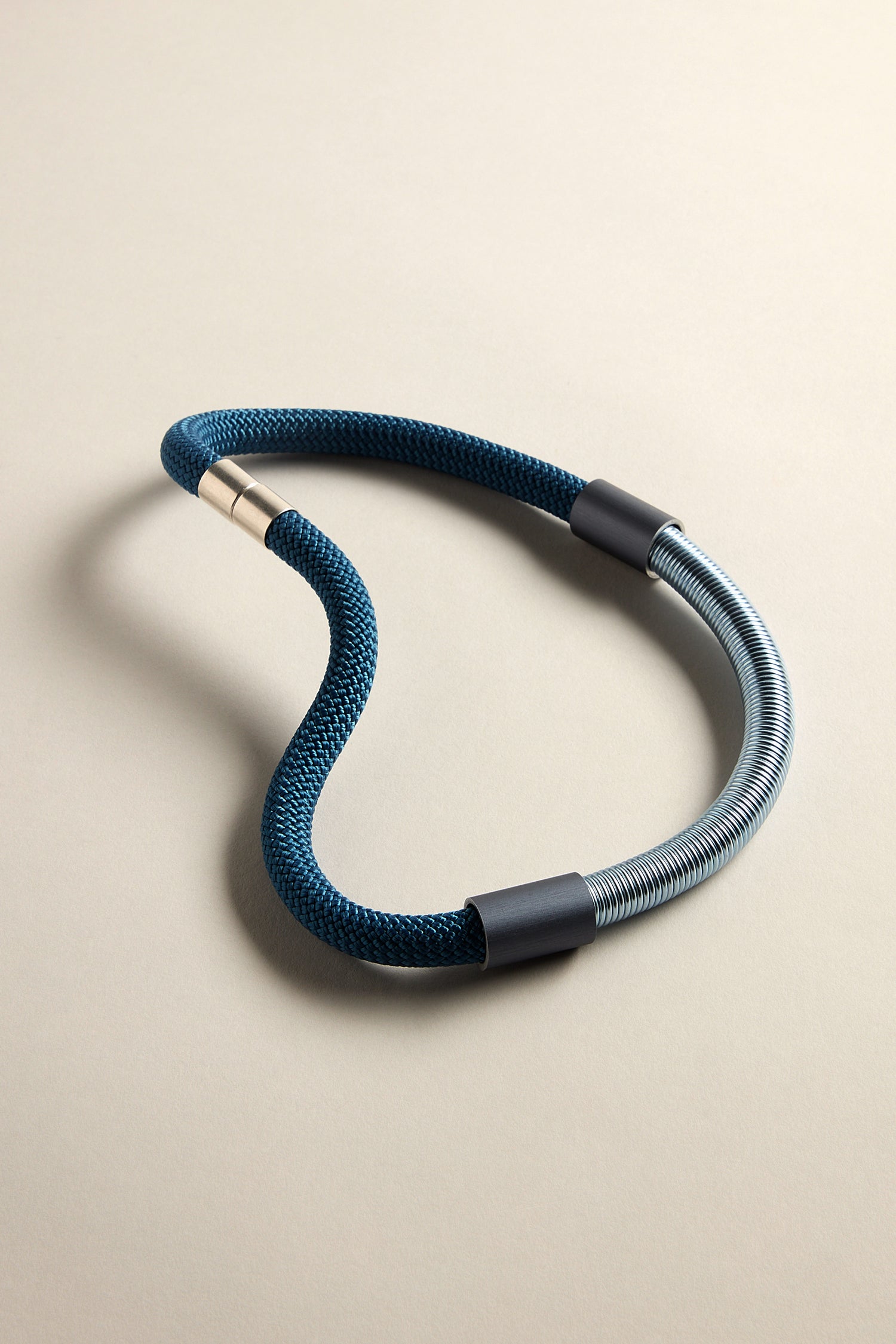 The Flow Cord Necklace, featuring a coiled design with dark blue and anodized aluminum segments, lies on a neutral-colored surface.