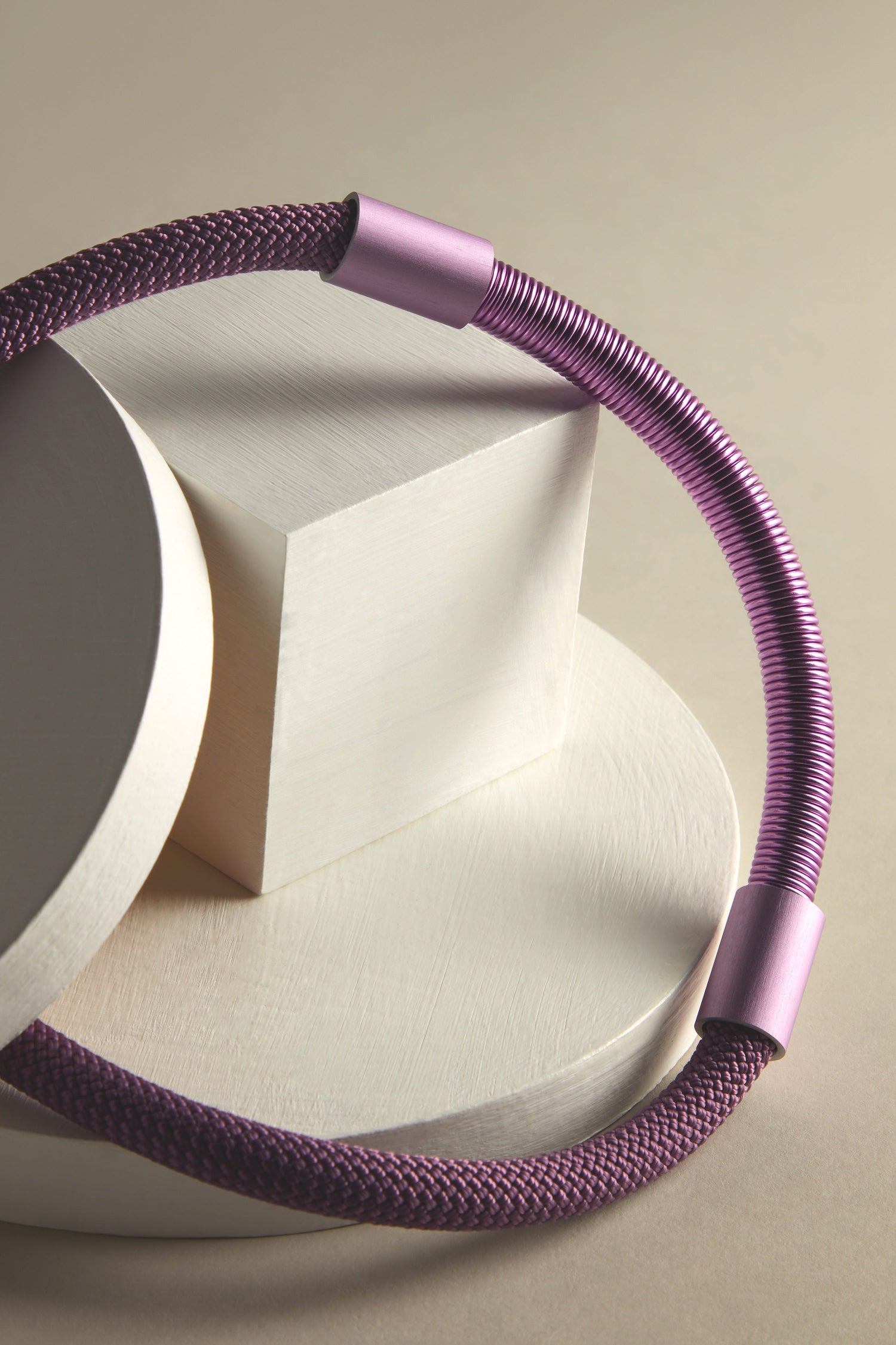 A purple, flexible cable with anodized aluminum connectors coils seamlessly on geometric white shapes, resembling a Flow Cord Necklace.