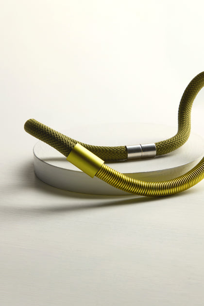 An Olive Flow Cord Necklace, resembling a yellow and green flexible hose, is skillfully arranged on a white surface.