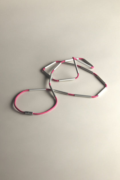 The Long Cylinders Necklace features a contemporary design with alternating pink cord and silver cylindrical beads, creating a striking statement against a neutral backdrop.