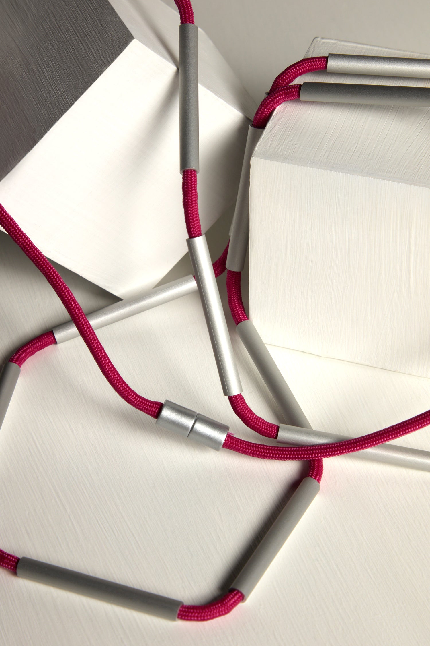 The Long Cylinders Necklace features a contemporary geometric design with blocks and magenta rope interwoven with silver cylindrical segments, set against a neutral background.