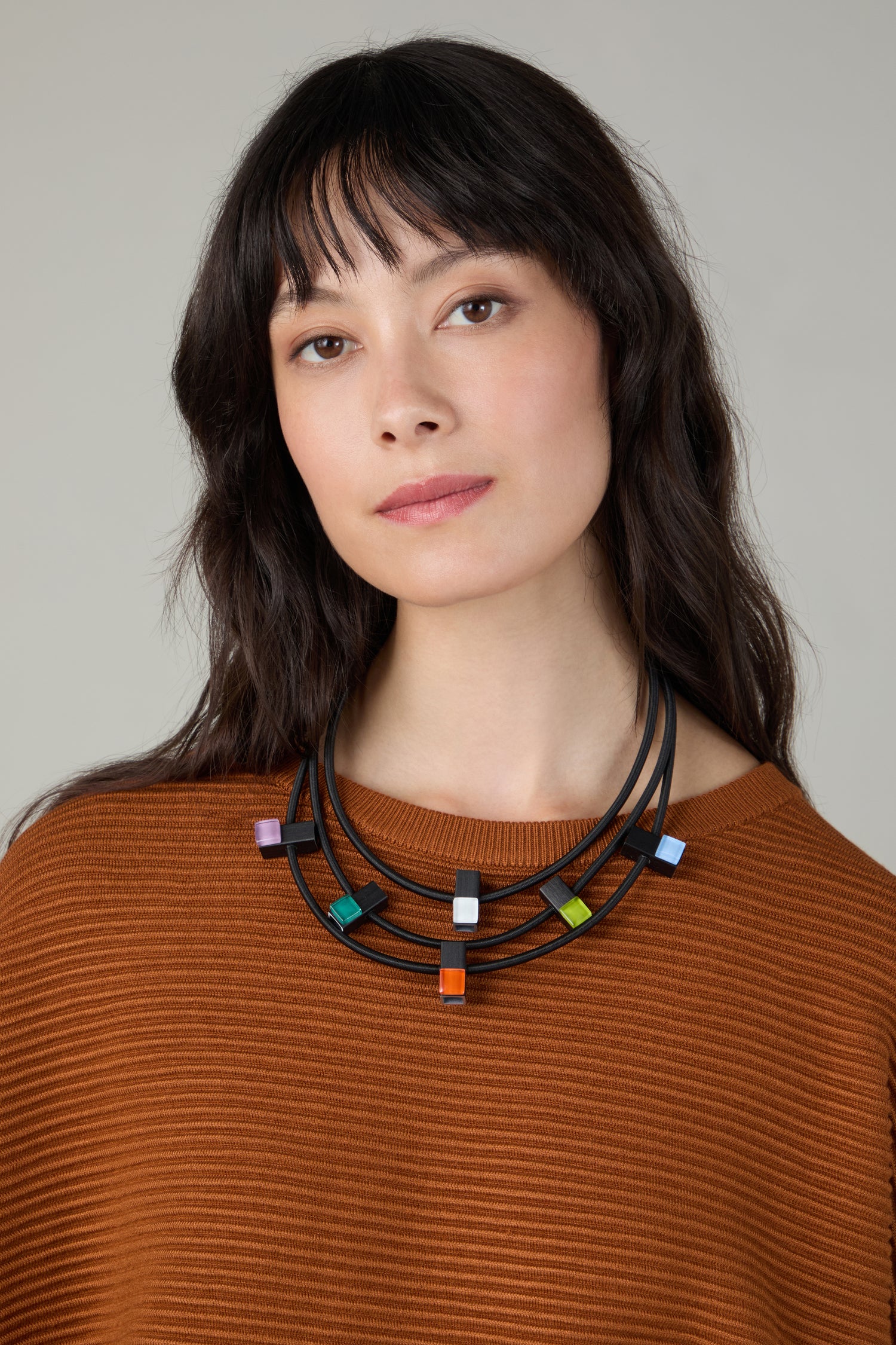 A woman with long dark hair wears a rust-colored sweater and the Colour Pop Glass Beads Necklace, featuring bold colorful geometric beads, posed against a neutral background—showcasing striking statement jewellery.