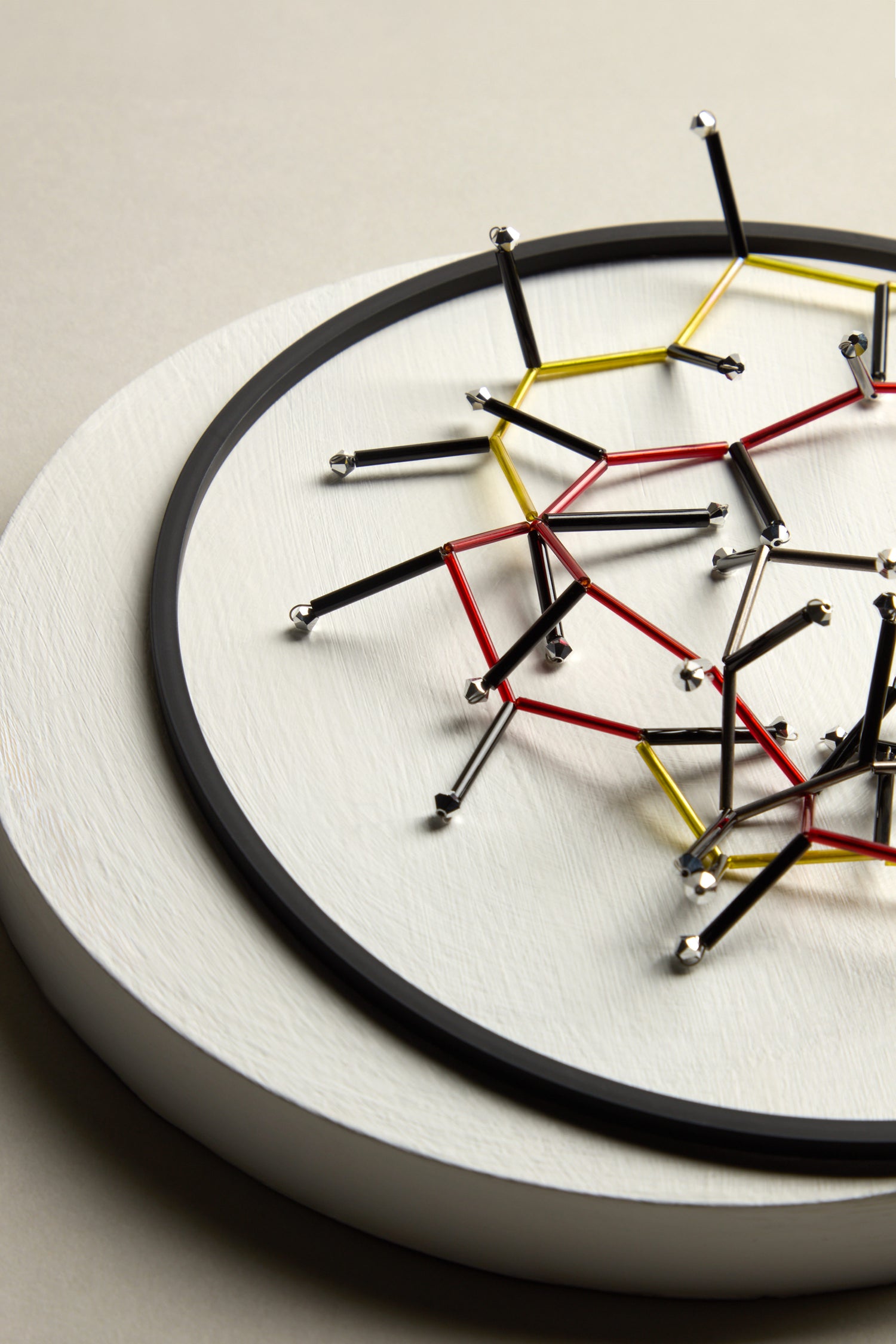 The Atomic Necklace is an avant-garde accessory with interlocking black, red, and yellow rods forming a geometric pattern on a round white base.