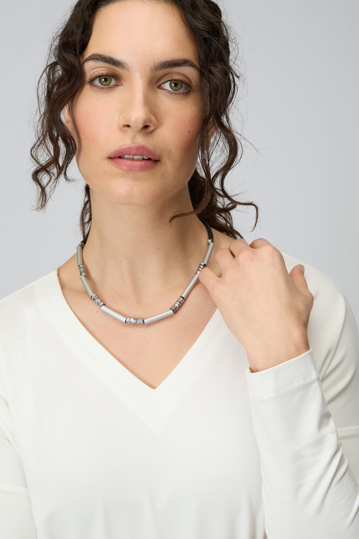 Woman with wavy dark hair in a white V-neck top wears the Silver Aluminium Hematite Necklace by Christina Brampti, gazing at the camera as her hand touches the striking accessory.